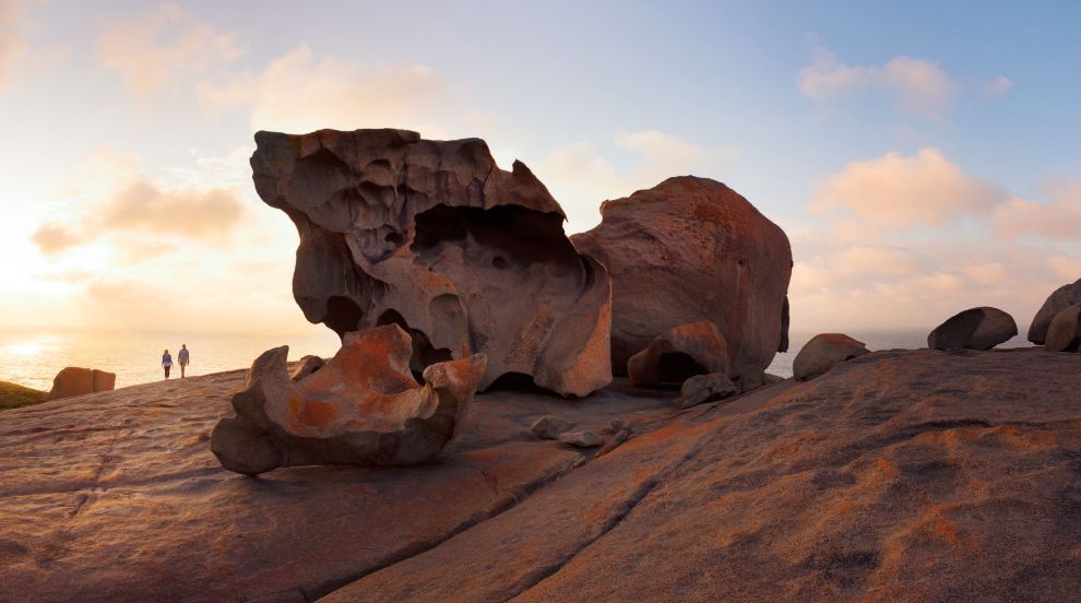 Rocky desert outcrop at sunset with two tiny hikers in the distance under a pink sky