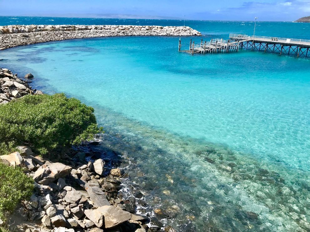 Tropical turquoise lagoon with rocky shore, breakwater, and wooden pier under a clear blue sky