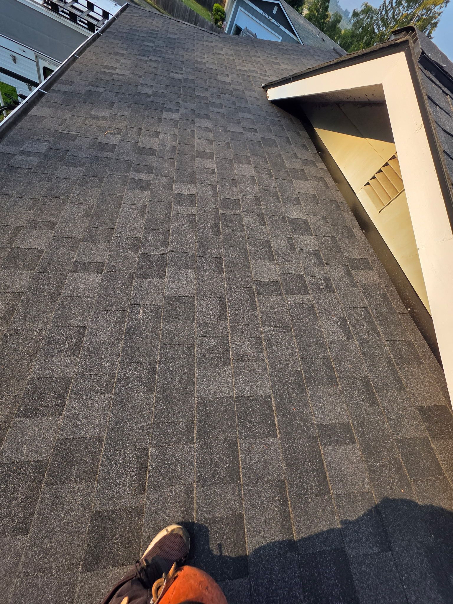 A high-angle view looking down a shingled roof towards a dormer, with a person's work boots visible at the bottom.