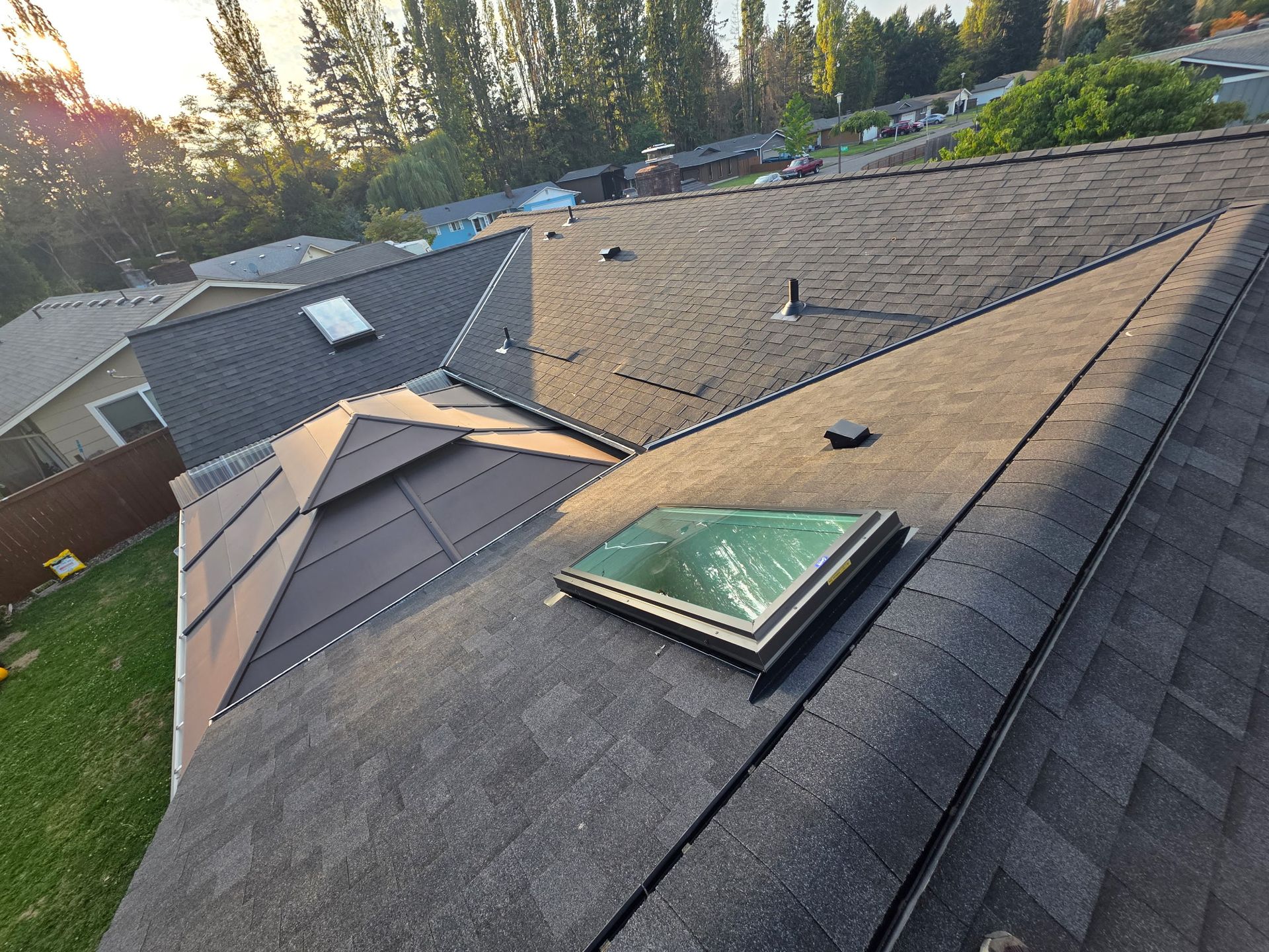 An aerial view of a dark shingled residential roof with two skylights and a metallic section, set on a sunny day.