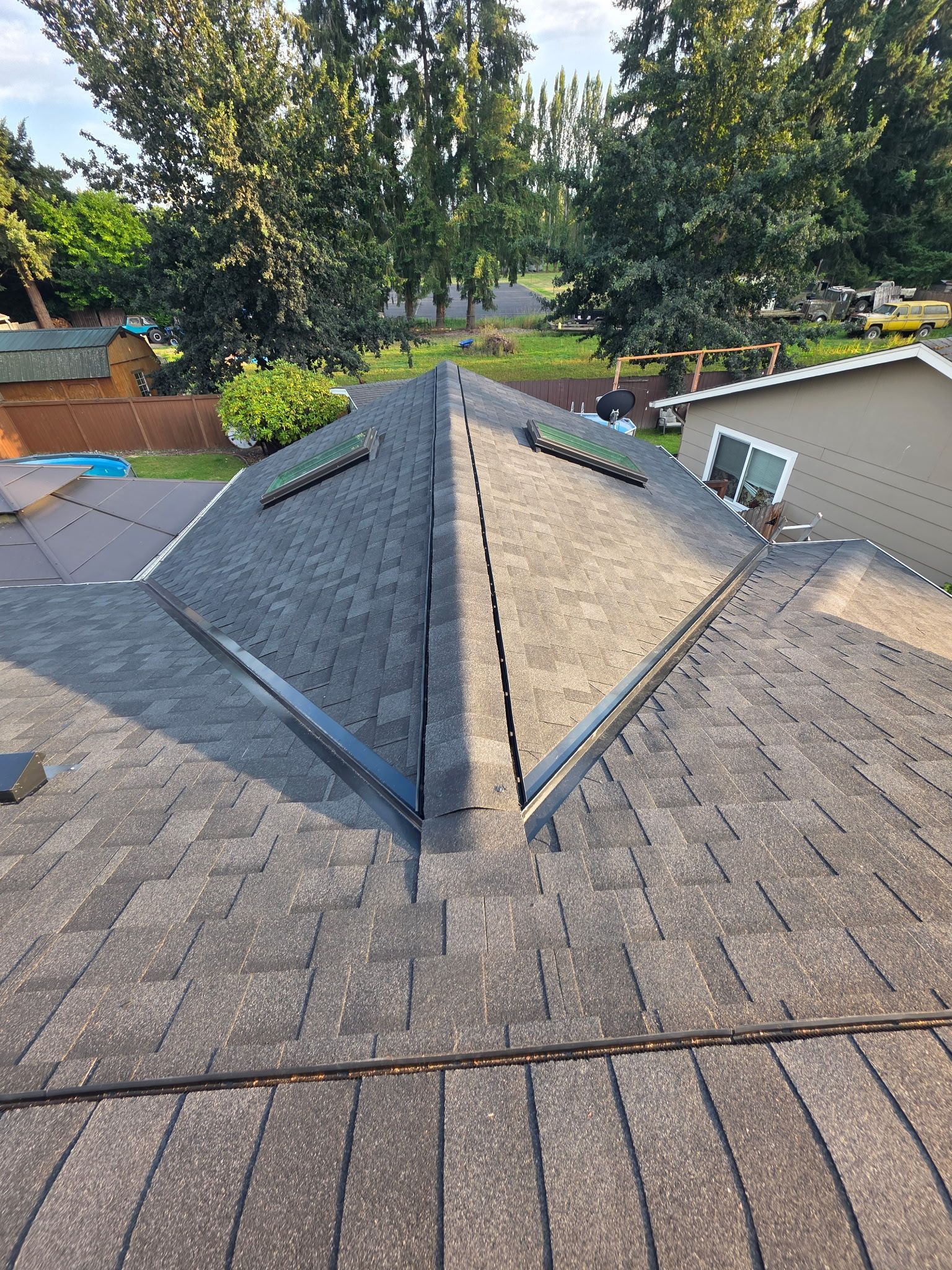 A view looking down at a gray asphalt shingle roof with two skylights and metal flashing on a sunny day.
