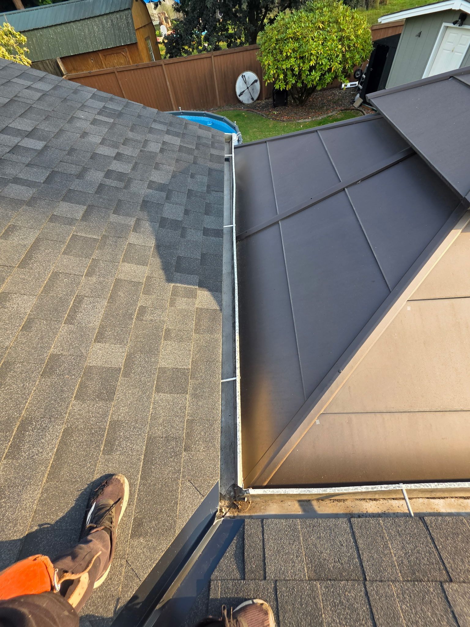 A person's feet stand on a shingled roof next to a metal roof section and a rain gutter in a residential backyard.