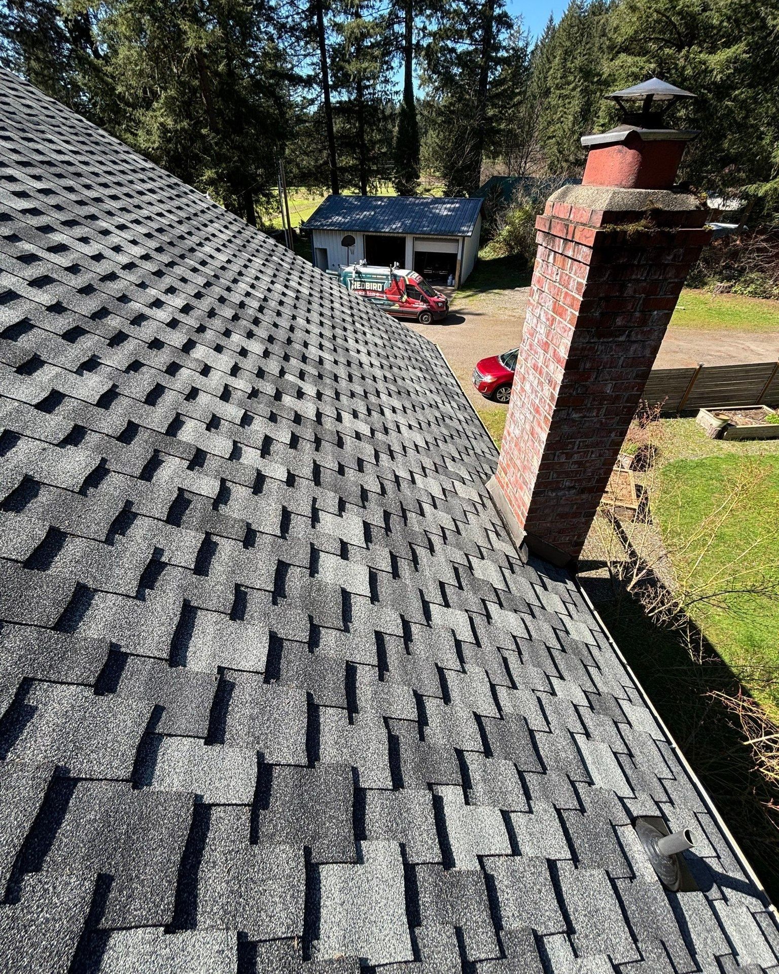 A high-angle view of a gray shingled roof sloping down toward a tall brick chimney, overlooking a yard and a small building.