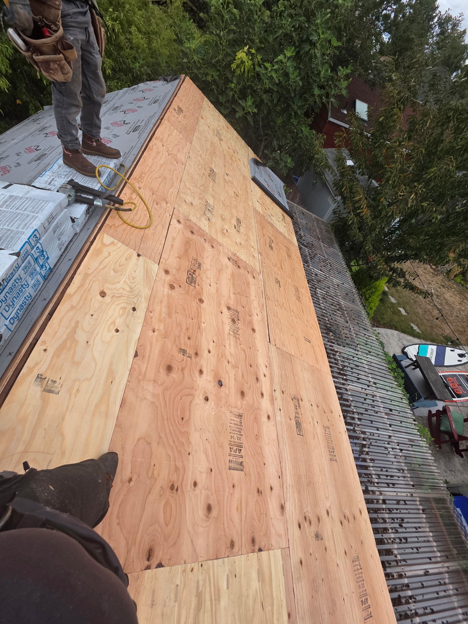 A worker stands on a partially installed plywood roof deck with underlayment and a shingled section visible nearby.