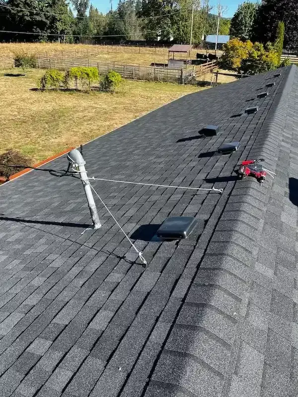 A gray shingled roof with a metal vent pipe supported by guy wires and several black rectangular roof vents.