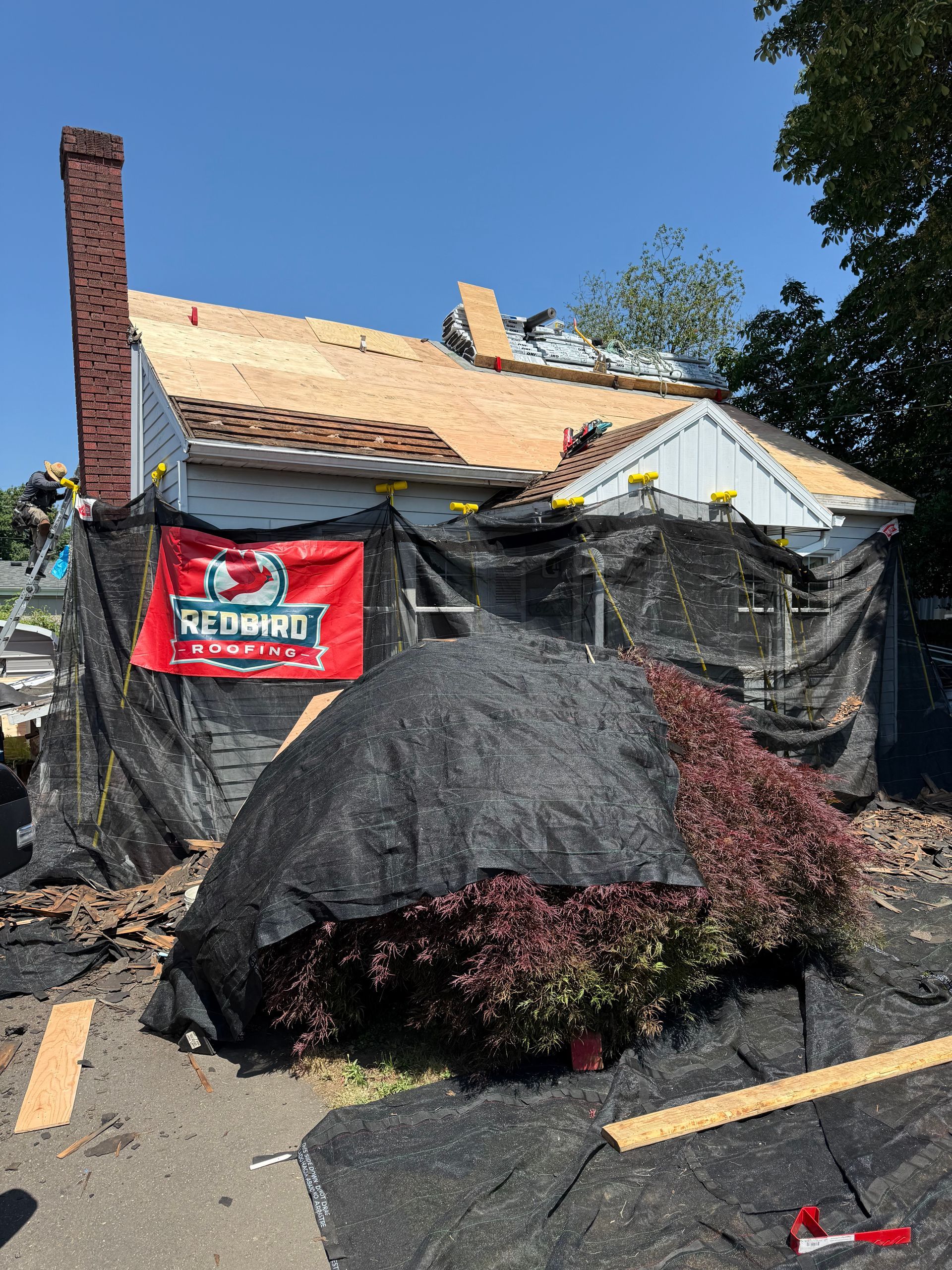 A house under construction with exposed roof sheathing, wrapped in protective black netting with a red sign attached.