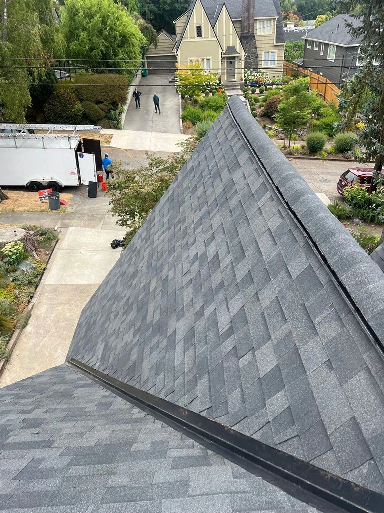A high-angle view of a grey shingled roof under construction, overlooking a driveway with a white trailer and workers.