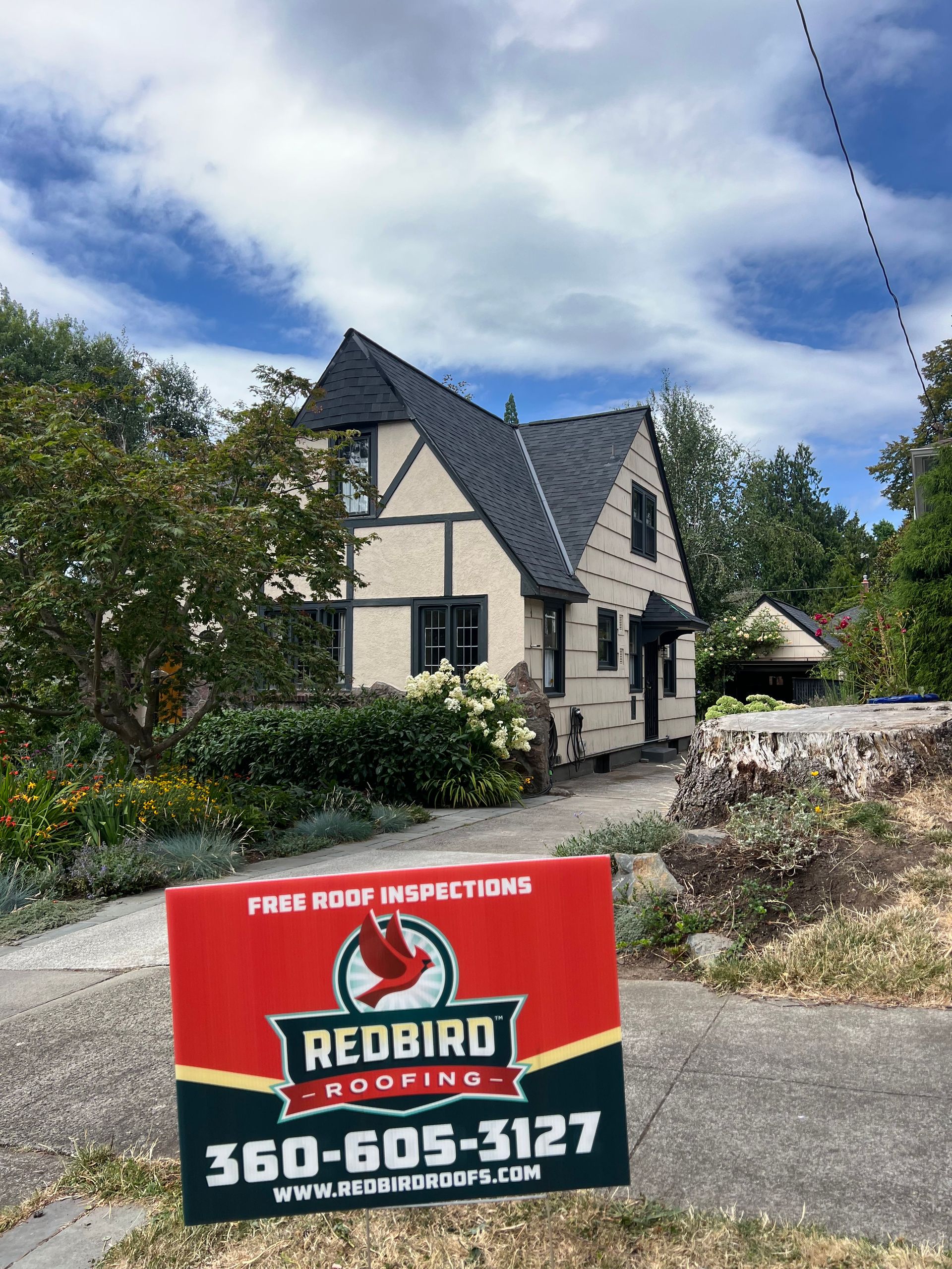 A Redbird Roofing sign stands in the foreground, with a tudor-style house and a clear blue sky in the background.