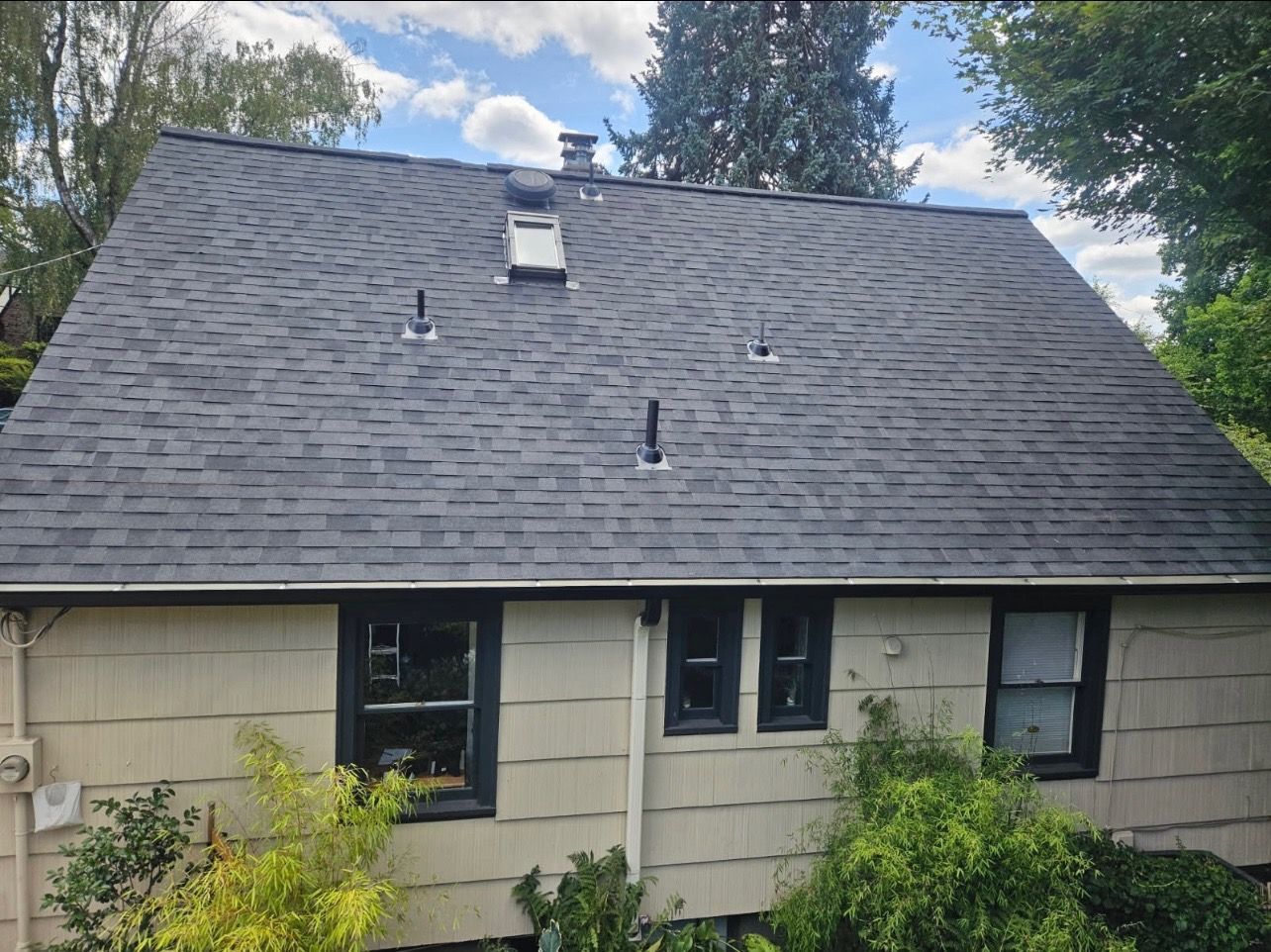 A tan one-story house with a grey shingled roof, dark window frames, and surrounding green foliage under a blue sky.