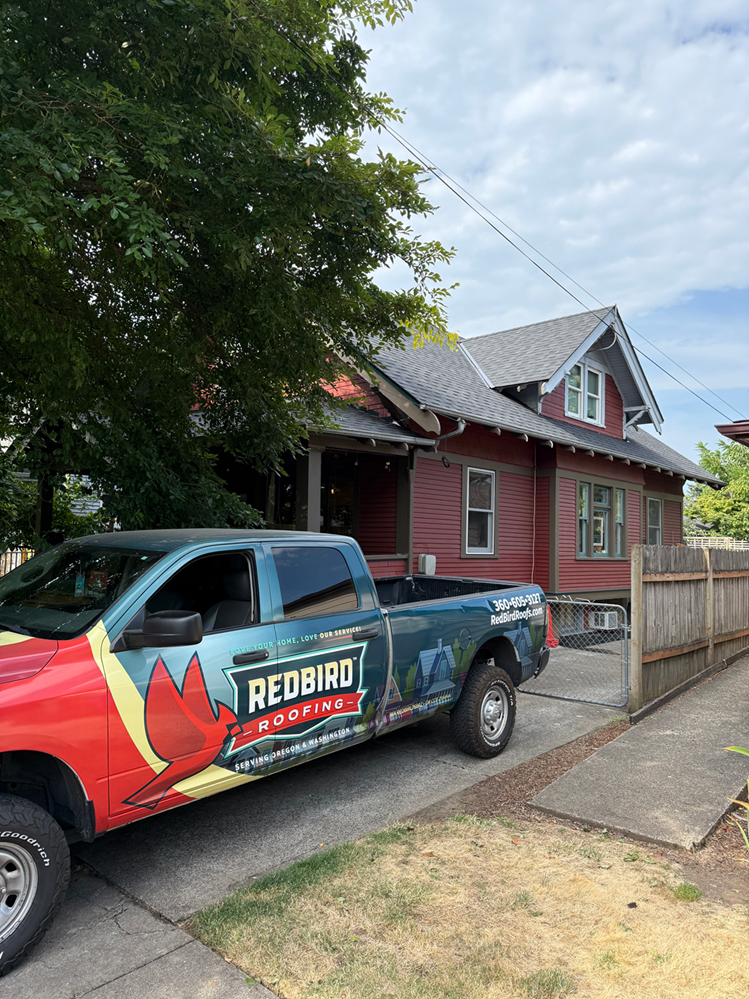 A red and blue Redbird Roofing truck parked in front of a red, one-story house on a sunny day.