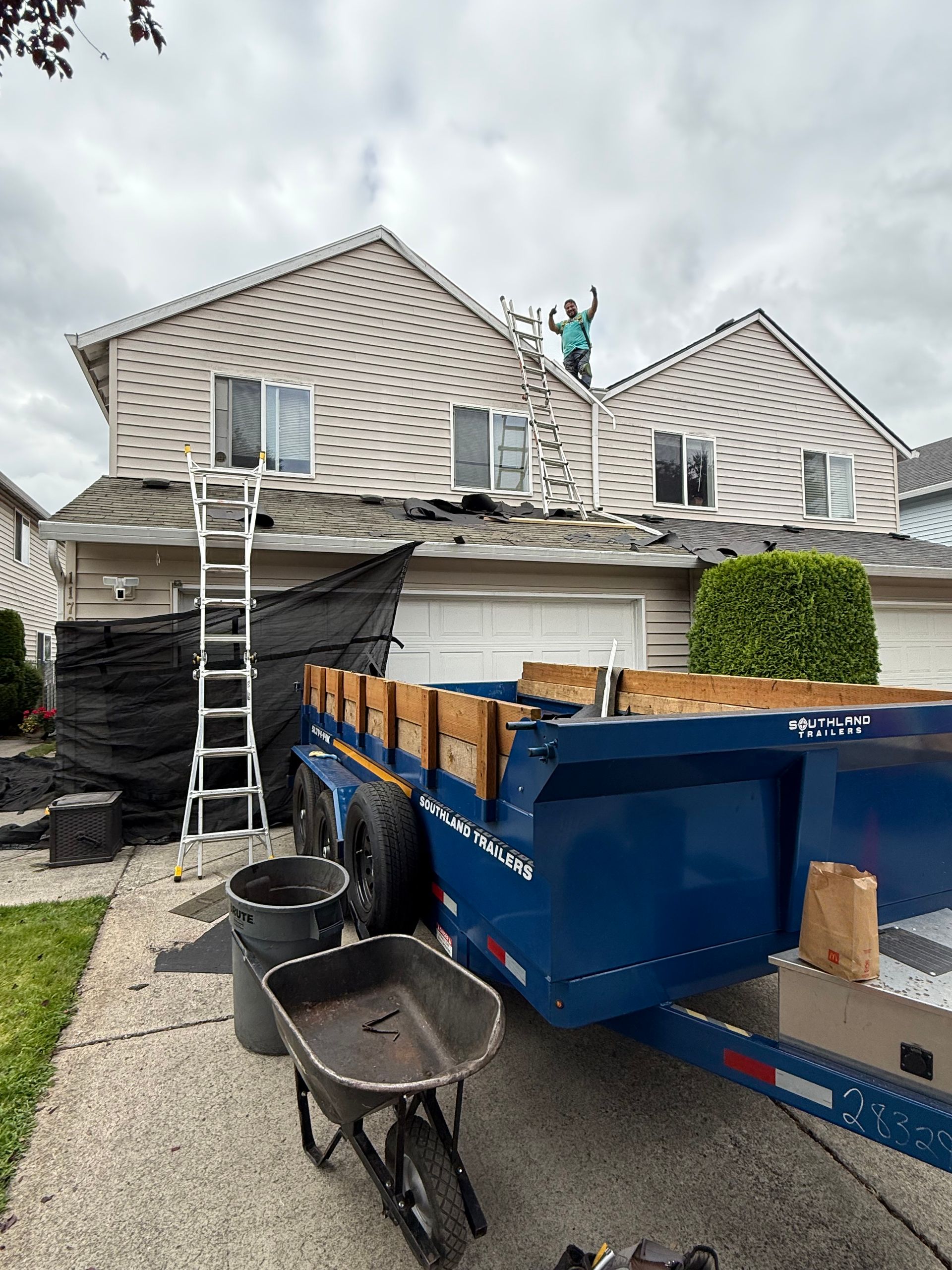 A person works on a house roof next to a ladder and a large blue dumpster parked in the driveway.