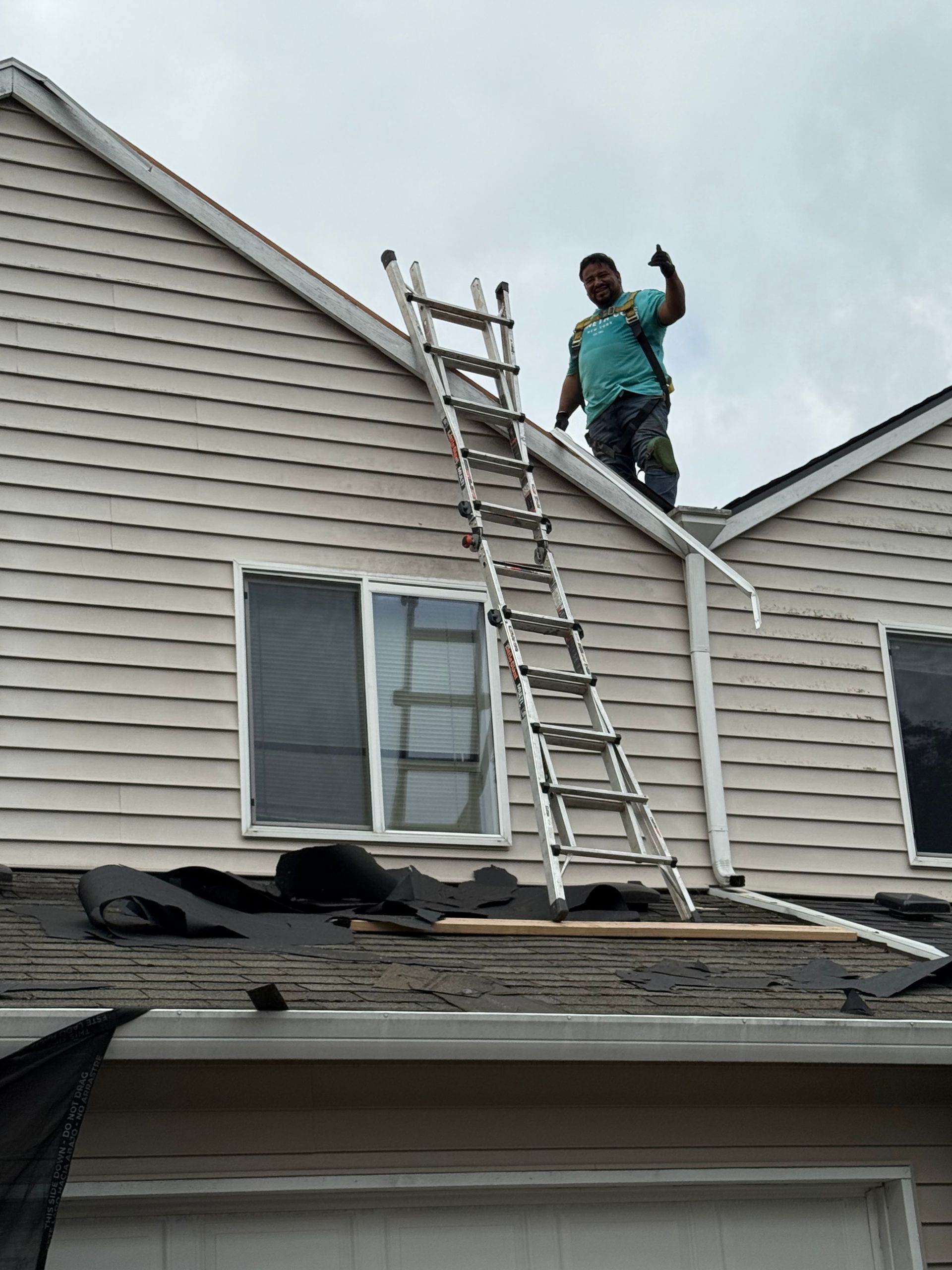A worker stands on a roof next to an extended ladder, giving a thumbs-up gesture against a residential siding exterior.