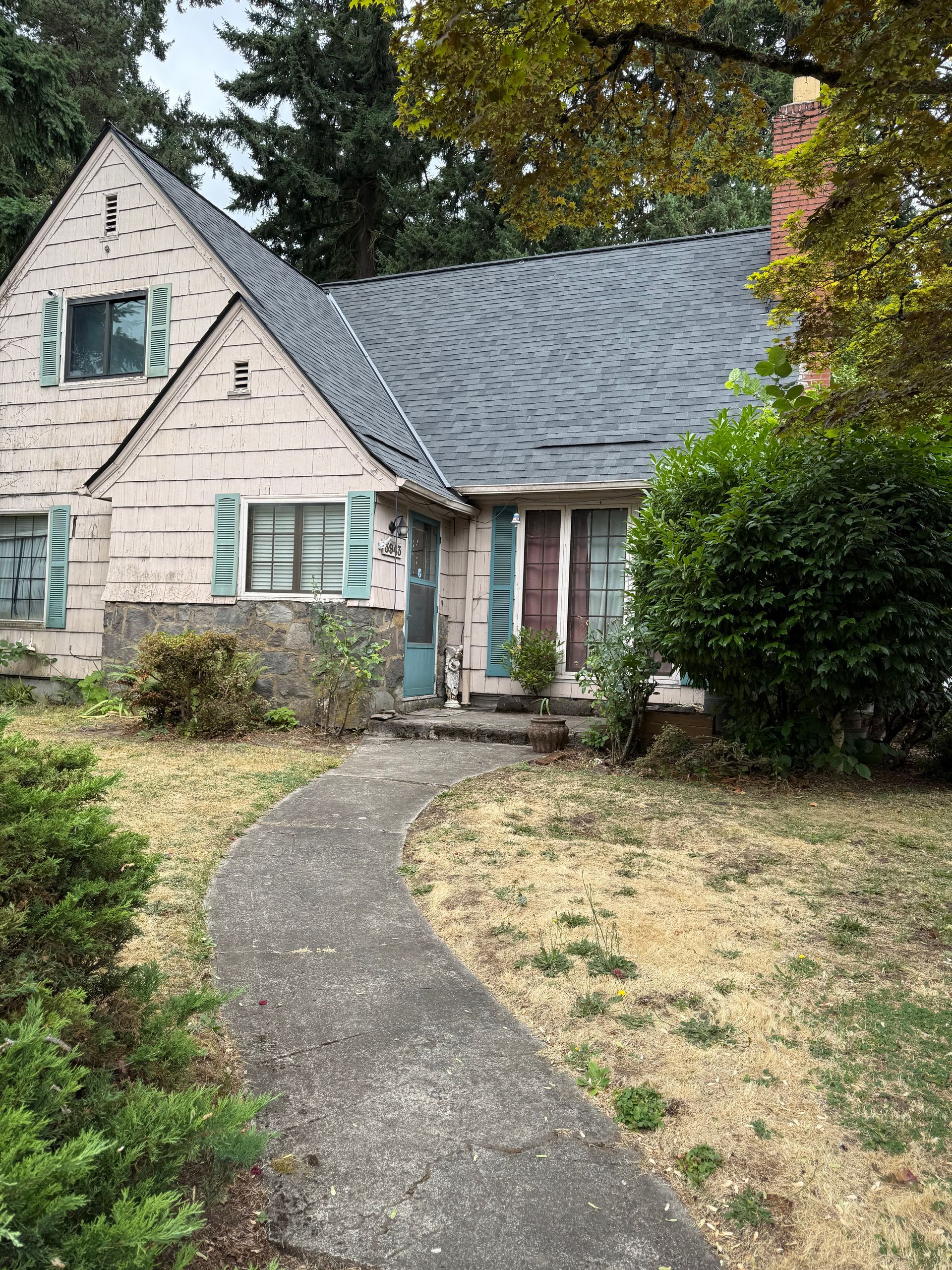 A two-story pink Tudor-style house with a gray roof and stone foundation sits behind a concrete path and dry lawn.