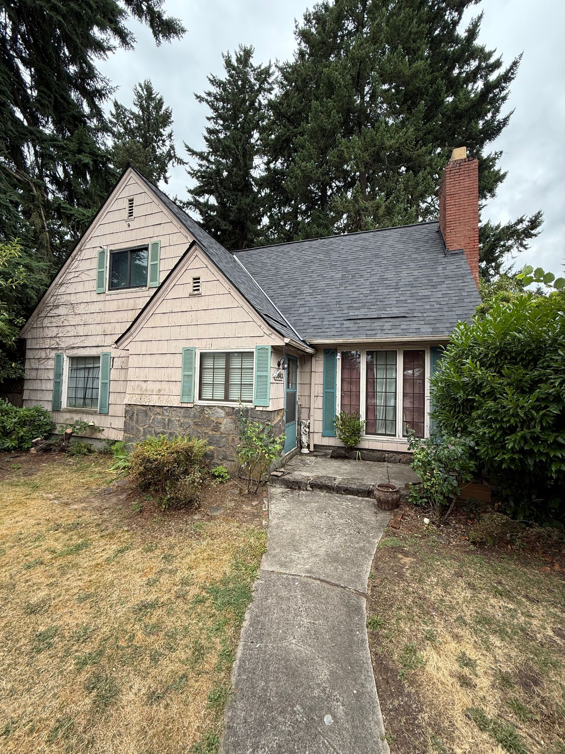 A two-story pink stucco house with green shutters, a slate roof, a spiral brick chimney, and a concrete front walkway.