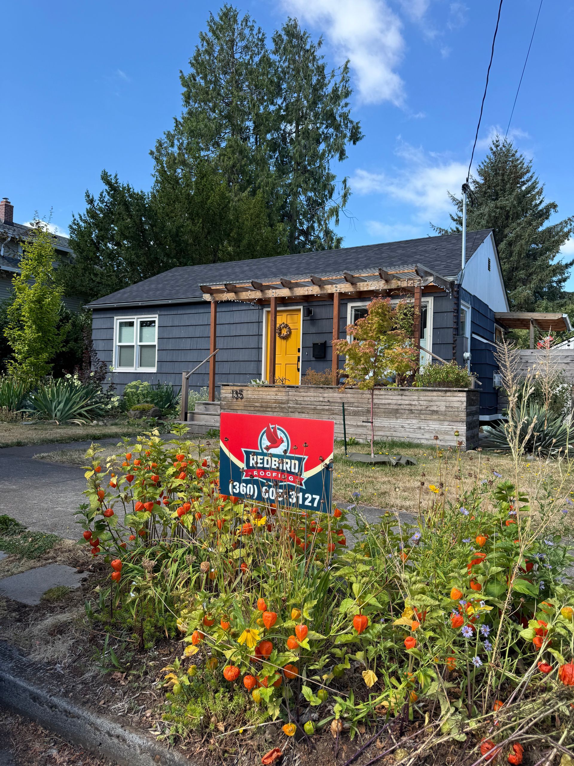 A grey house with a bright yellow front door, a wooden pergola, and a sign in the foreground amidst a garden.