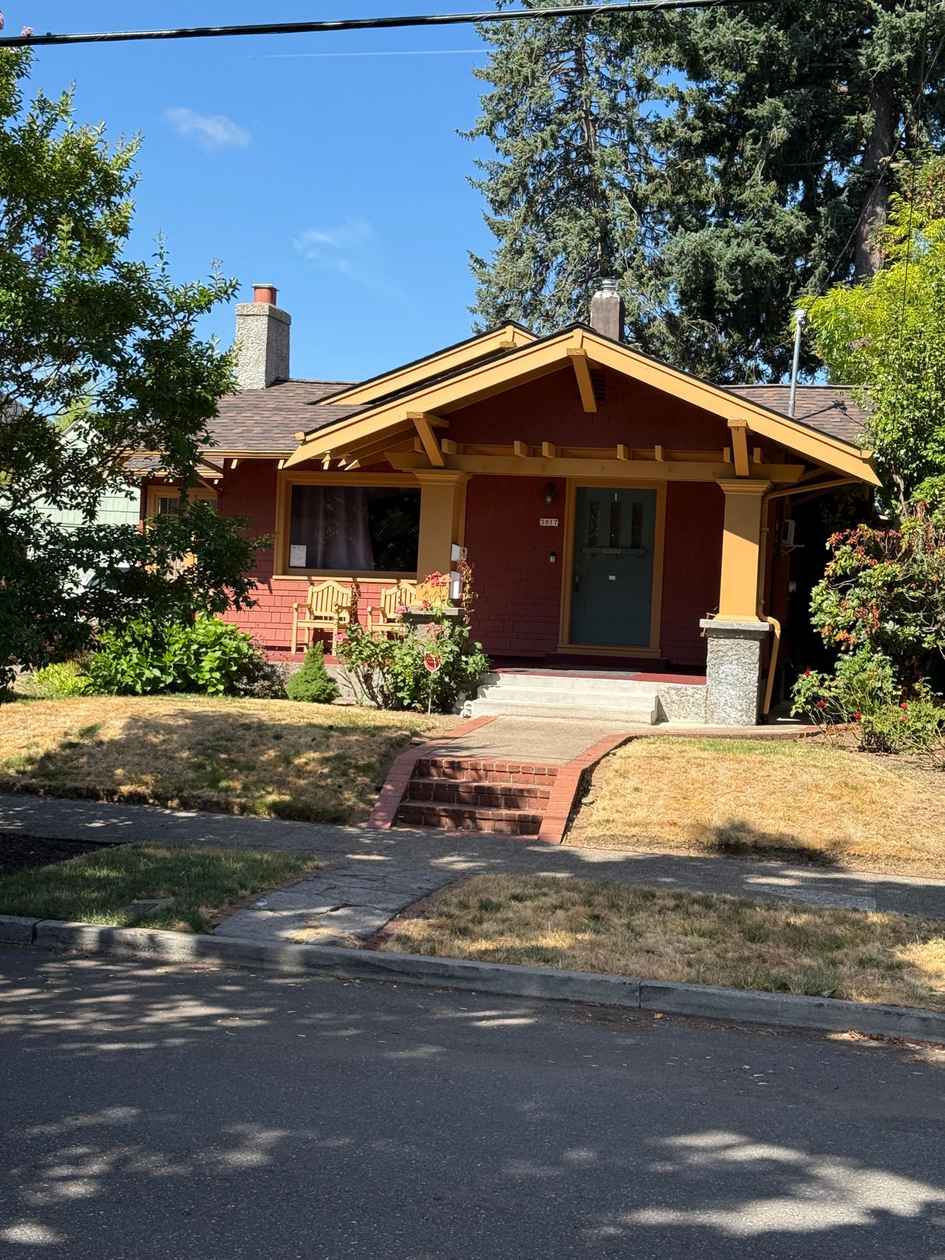A red bungalow with a stone-based front porch and roof trim, viewed from the street on a sunny day with a dry front lawn.
