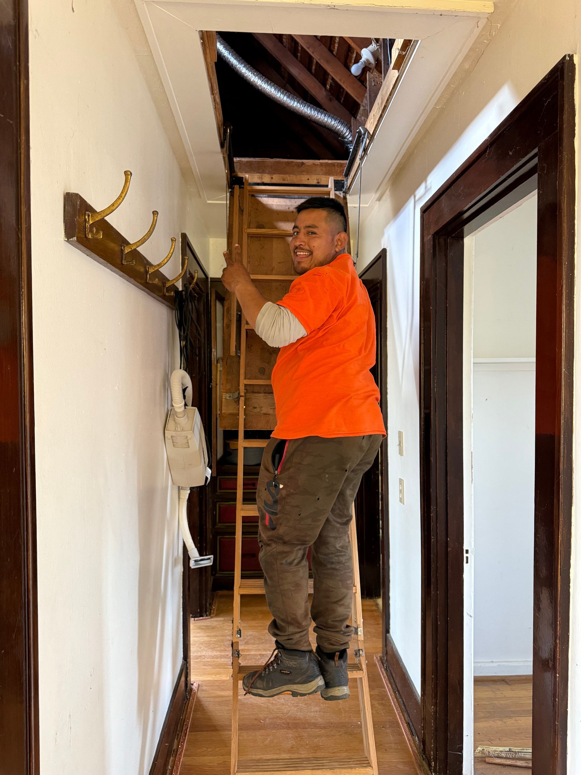 A person in an orange shirt climbs a wooden ladder into an attic opening in a hallway with dark wood trim and wall hooks.