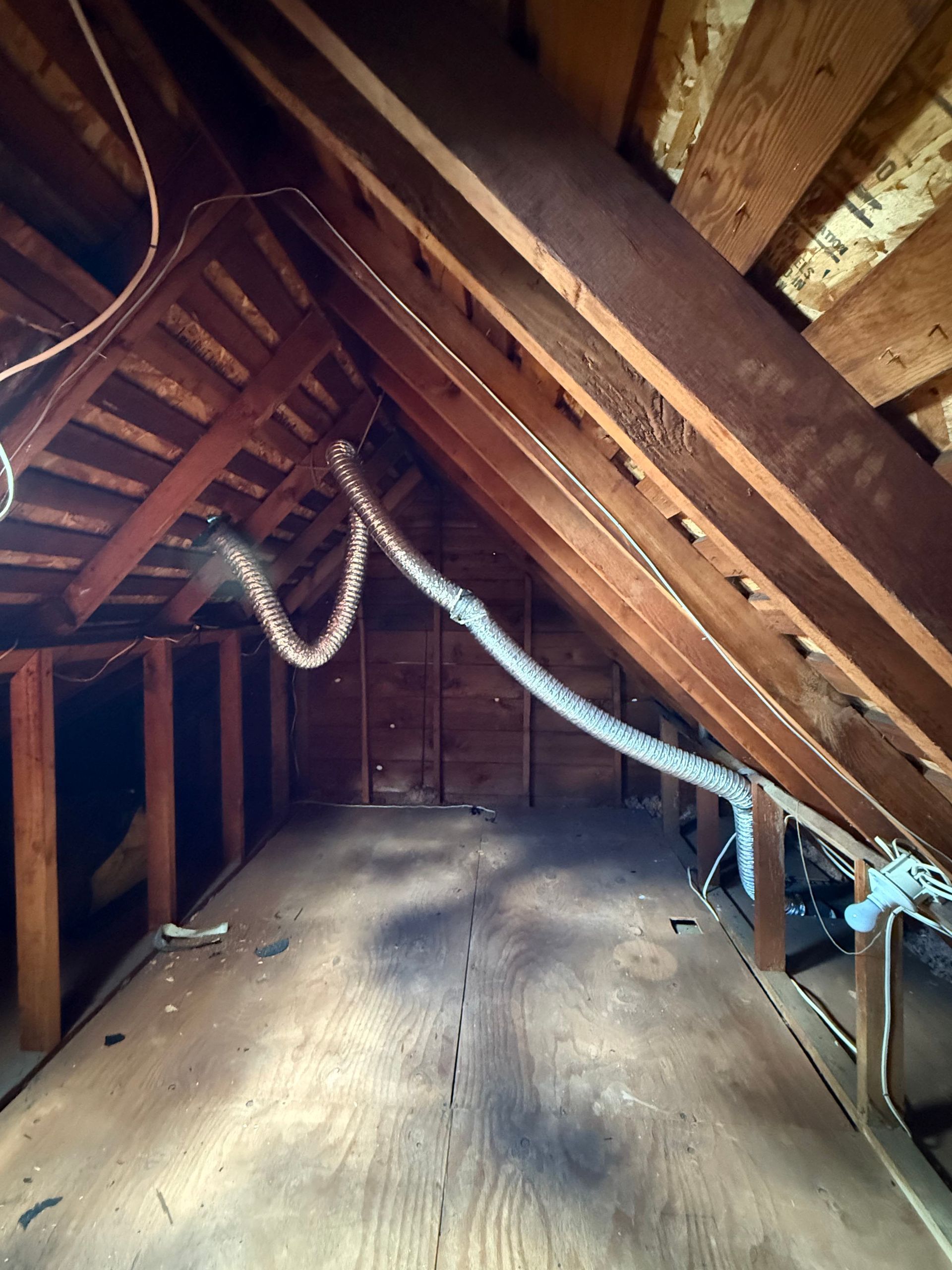 An attic interior showing exposed wooden rafters and a flexible metallic vent pipe running across the wooden floor.