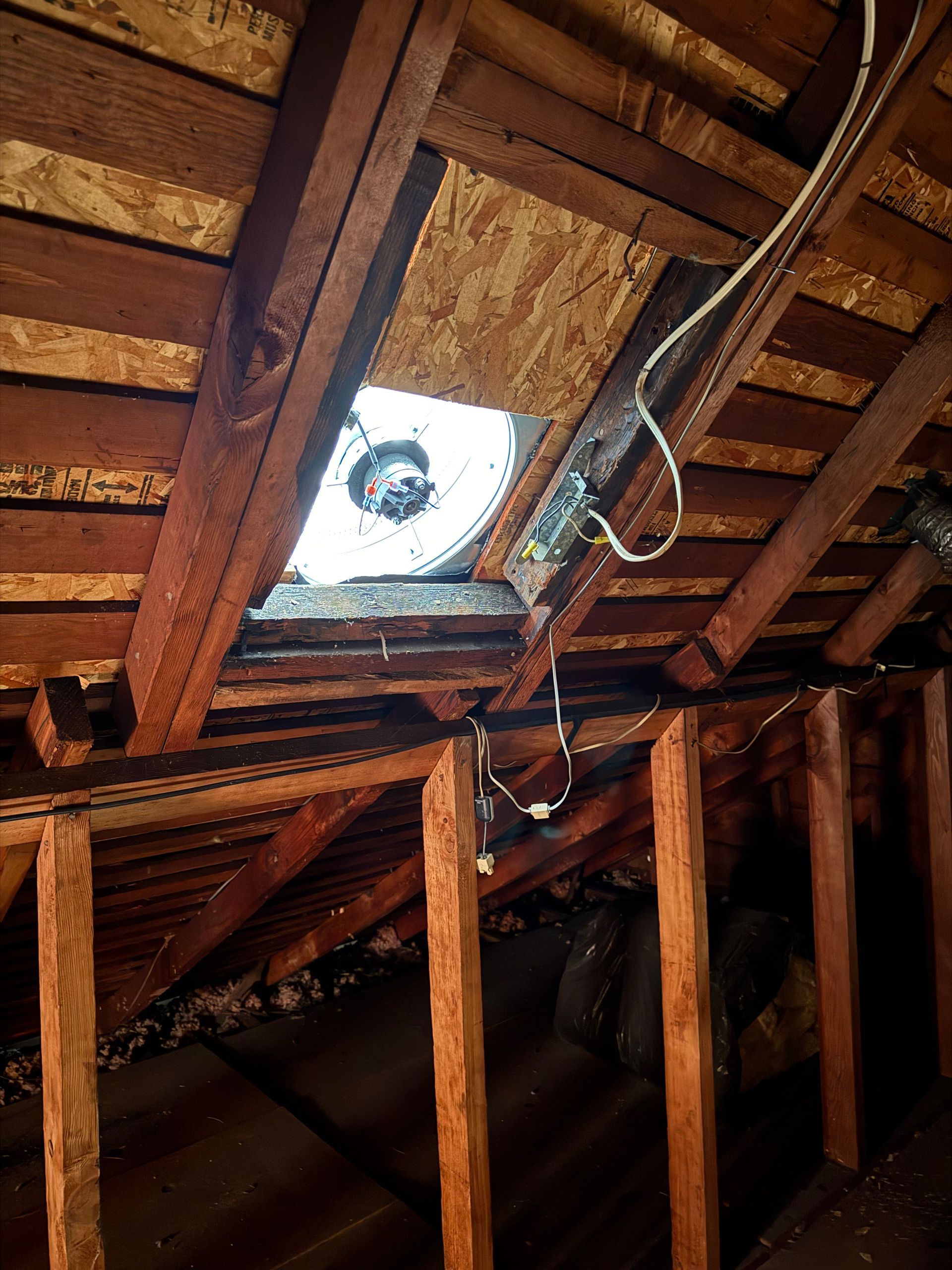 An attic interior showing wooden rafters, exposed framing, and a round ventilation fan installed in the roof decking.