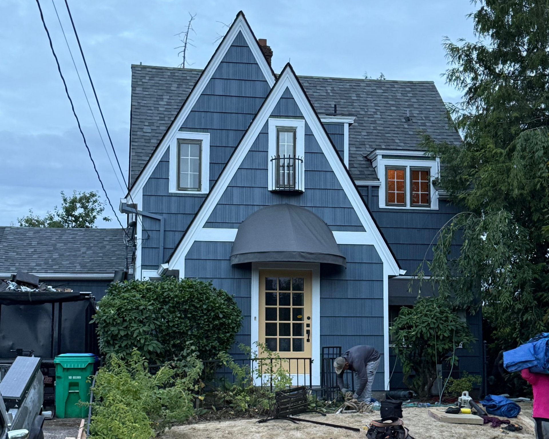 A blue two-story house with a prominent pointed roof, a yellow front door, and a person working in the front yard.