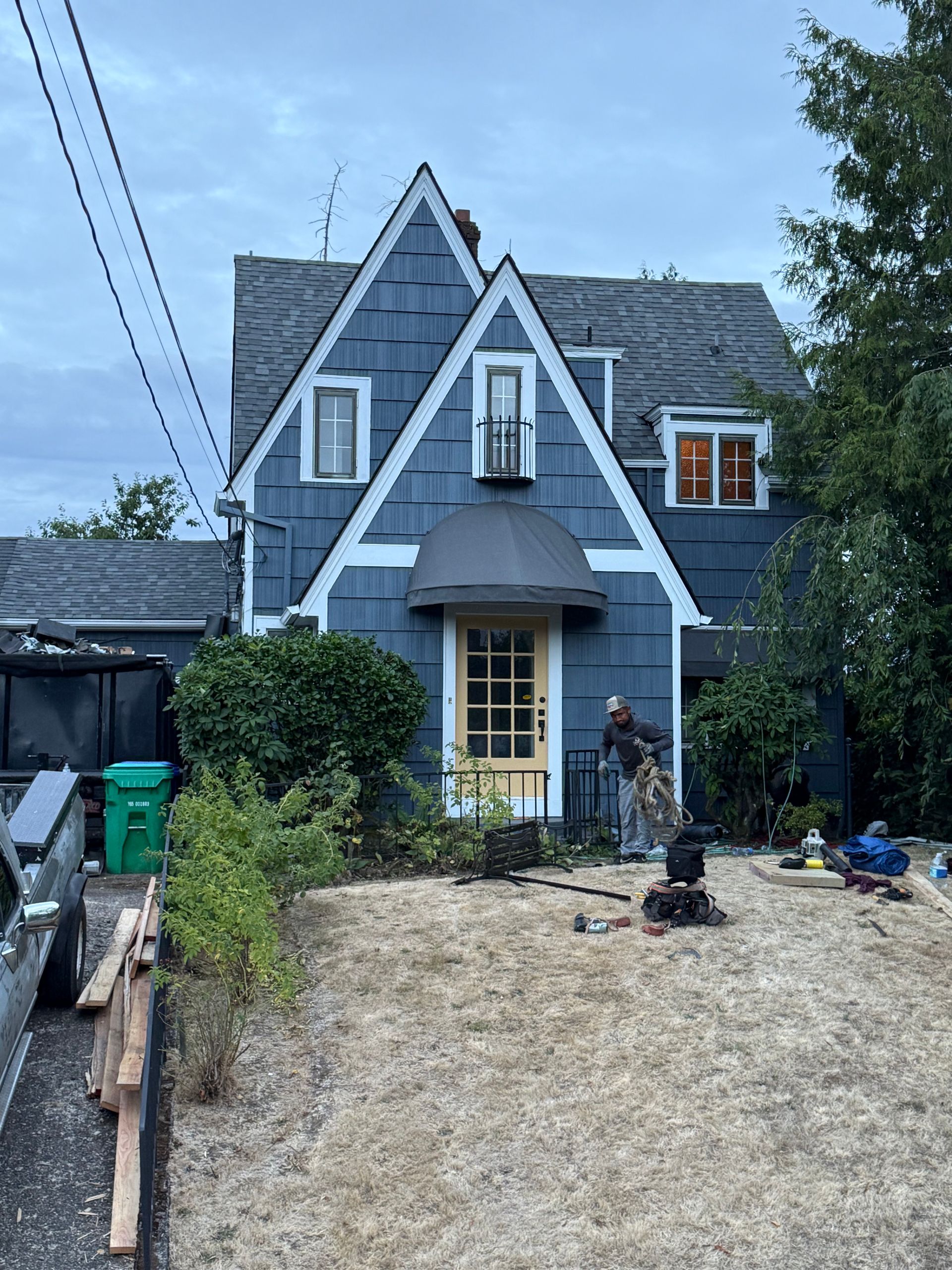 A blue, two-story house with a steep gable roof, a yellow front door, and an awning stands beside a sparse front yard.