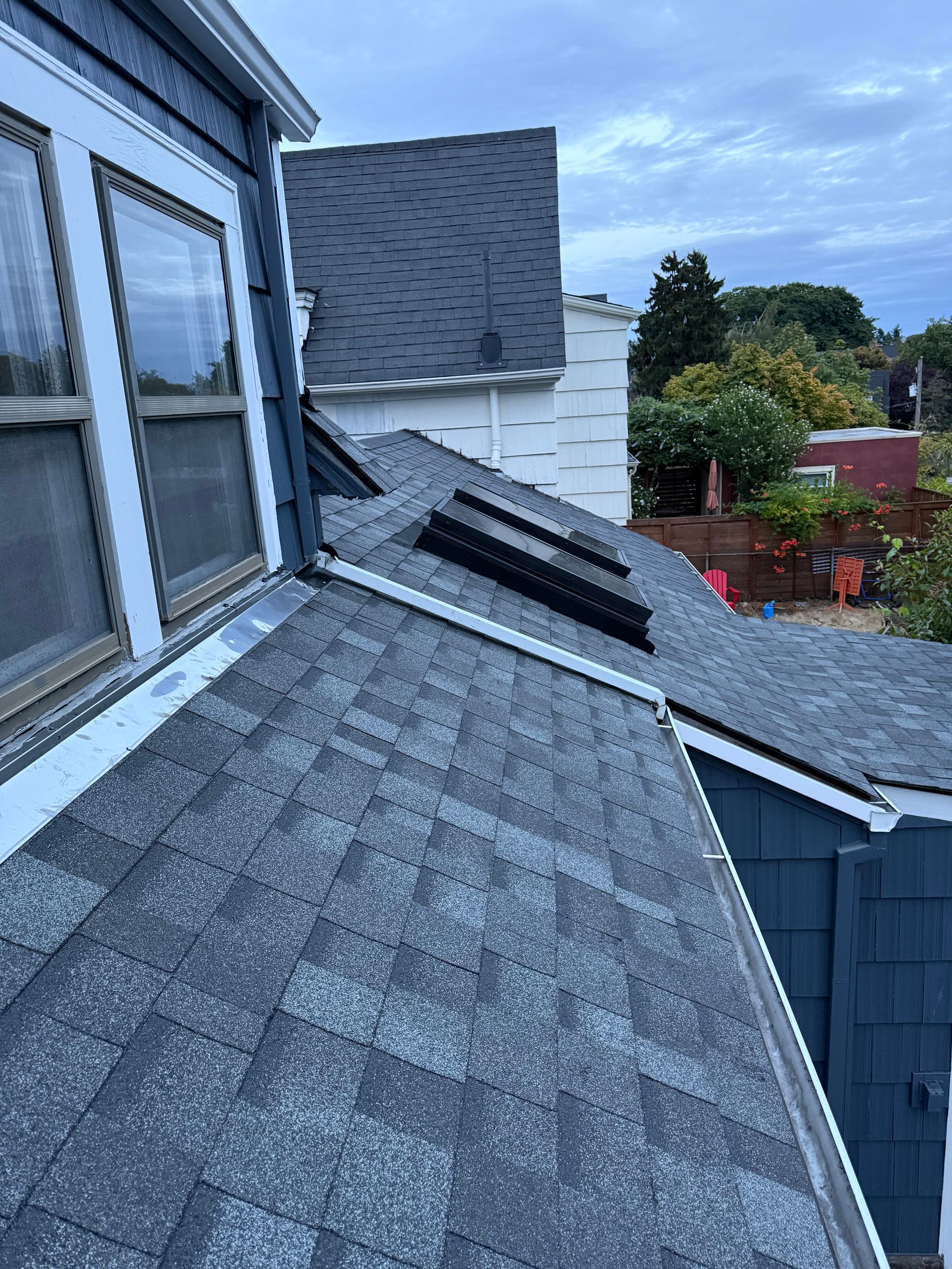 A high-angle view of a gray asphalt shingle roof with skylights, adjacent to a house with blue siding and white trim.