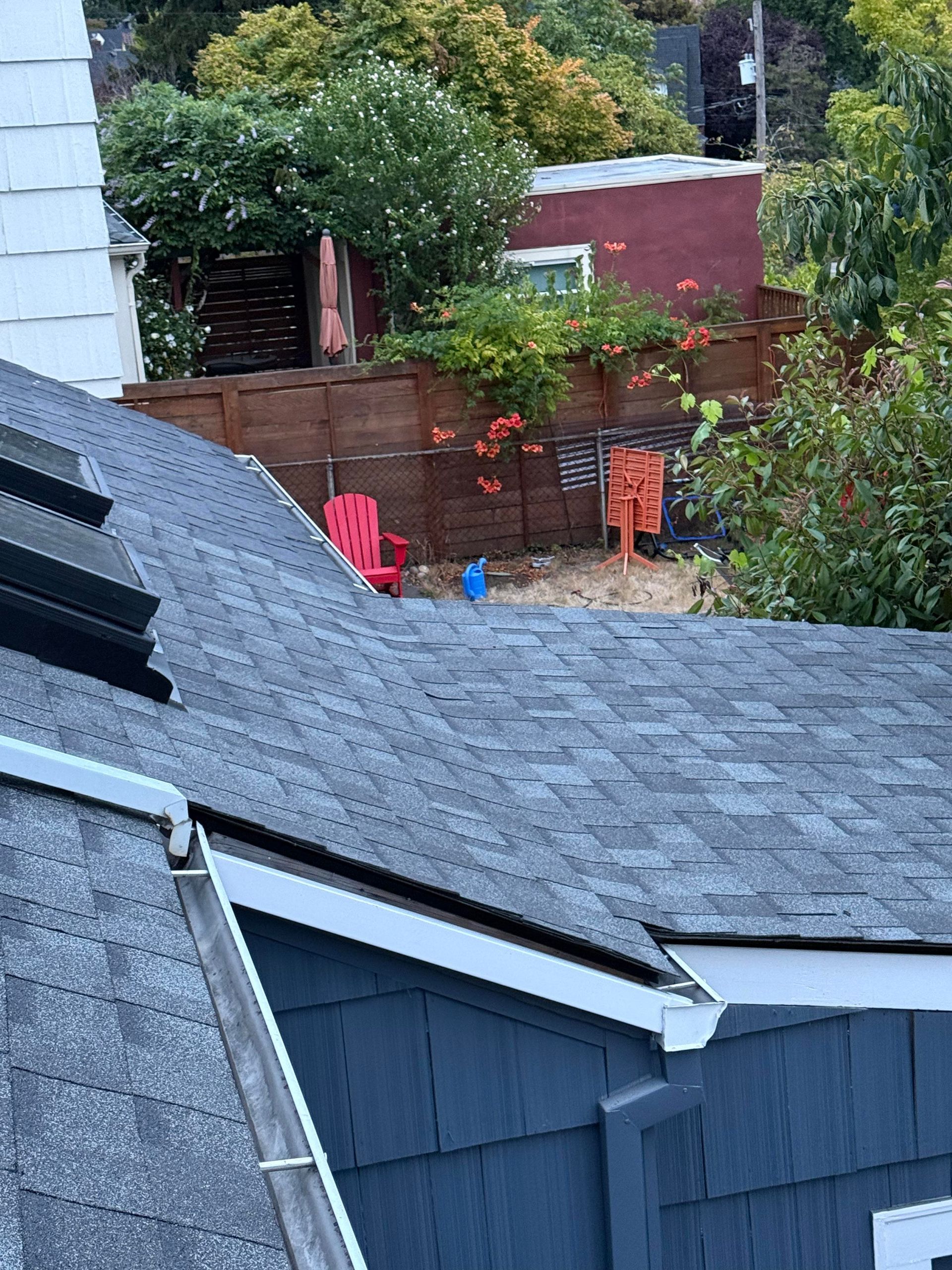 A view from a high vantage point overlooking a dark-shingled roof and a backyard with a wooden fence, plants, and red chair.