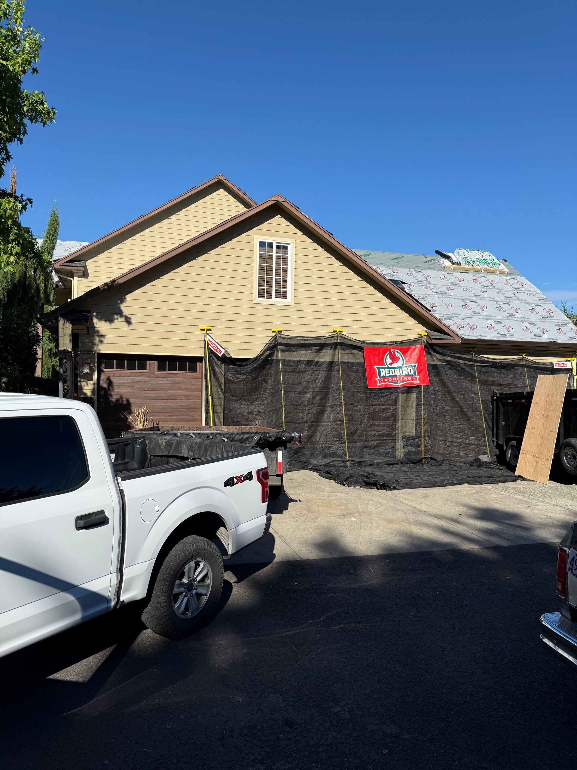 A white truck is parked in front of a house under construction with roof underlayment and black protective netting.