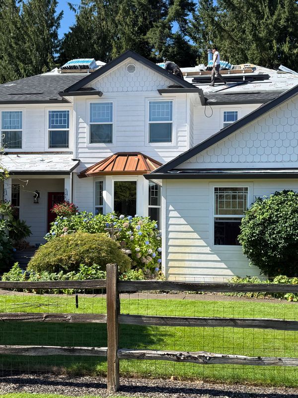 A two-story white house with a partially replaced roof, featuring a brown metal awning and a front yard with a wooden fence.