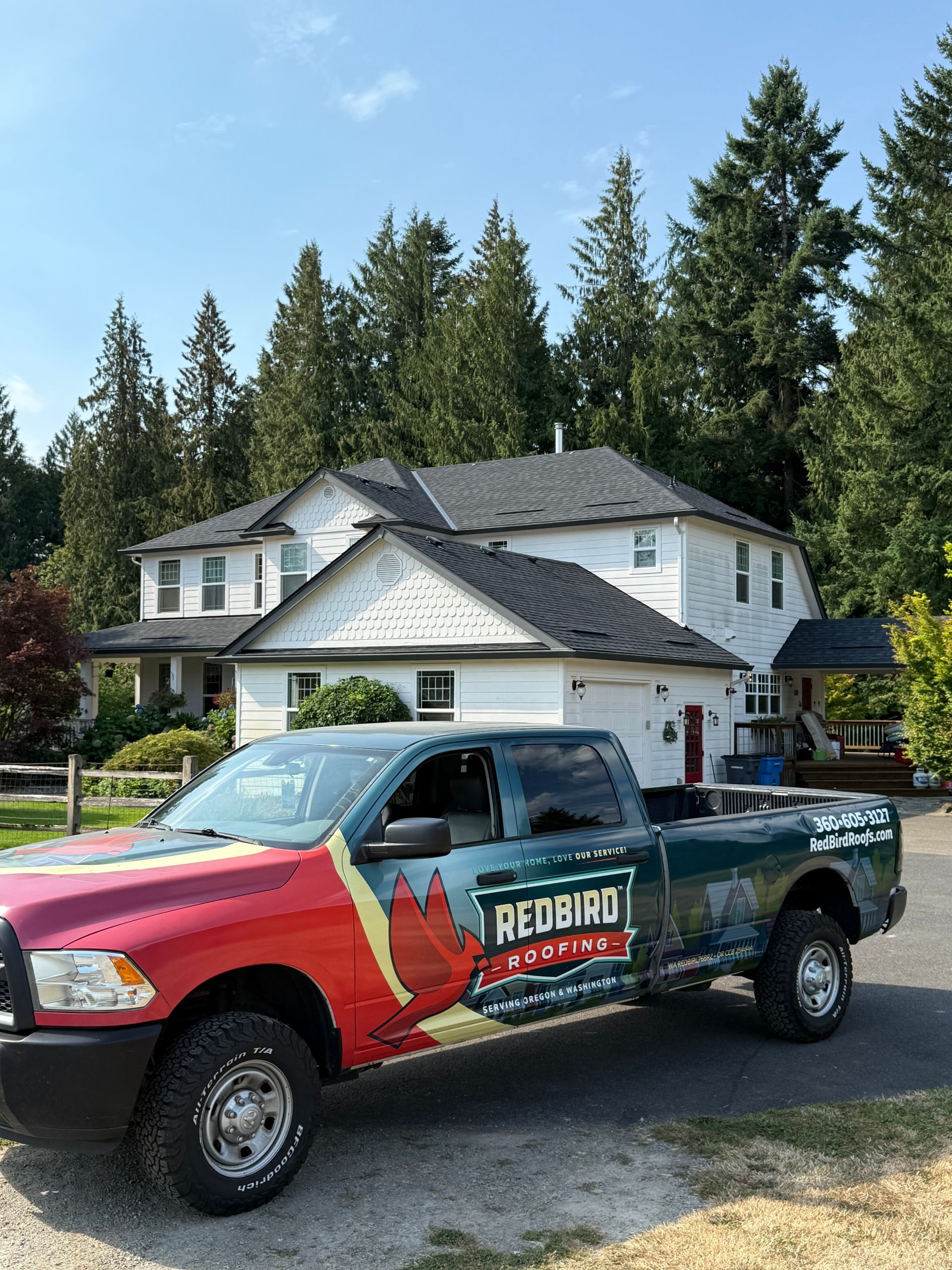 A red and green Redbird Roofing pickup truck parked on a gravel driveway in front of a white two-story house and trees.