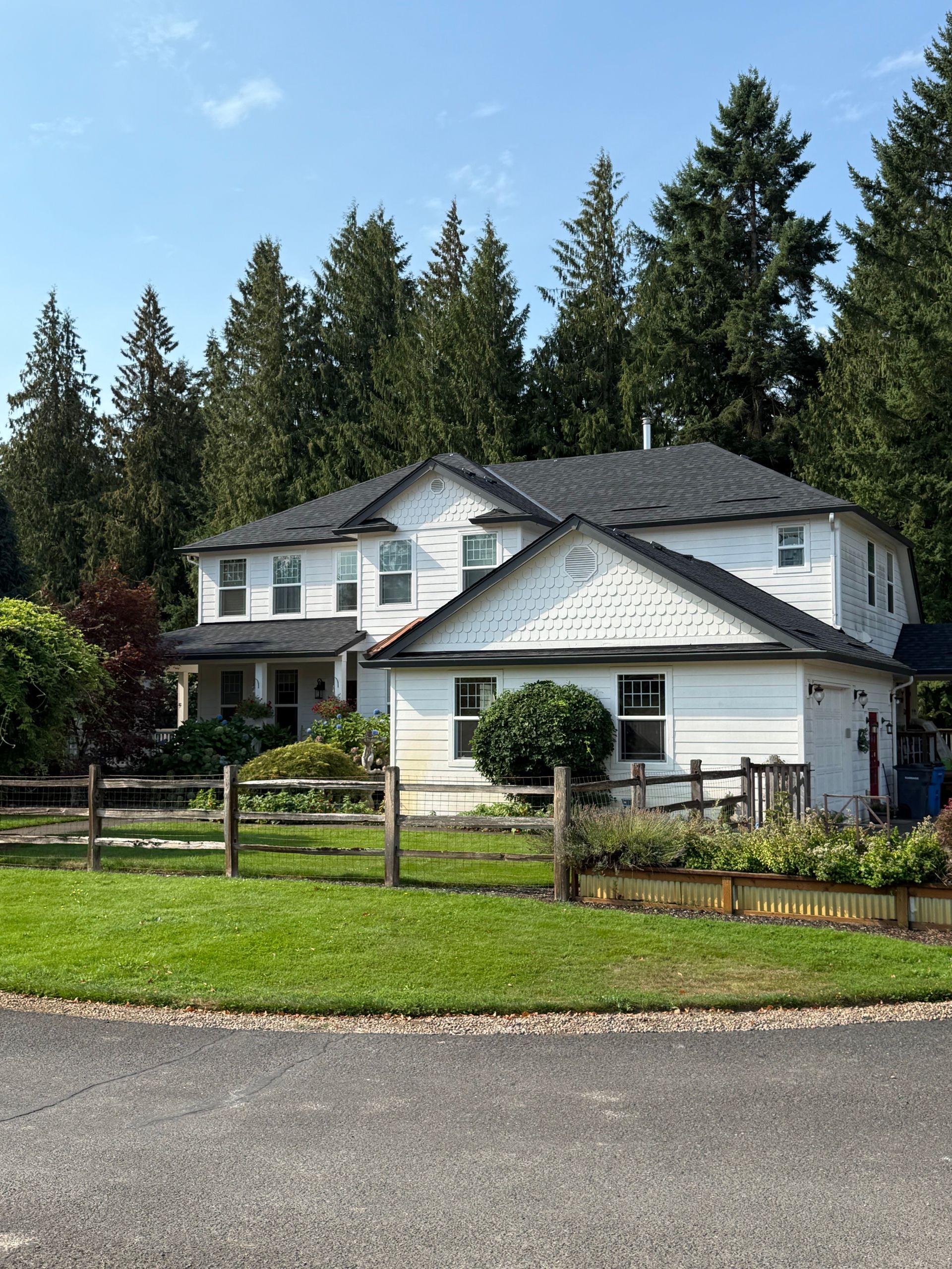 A two-story white house with a dark shingled roof, surrounded by a wooden fence, lawn, and dense forest background.
