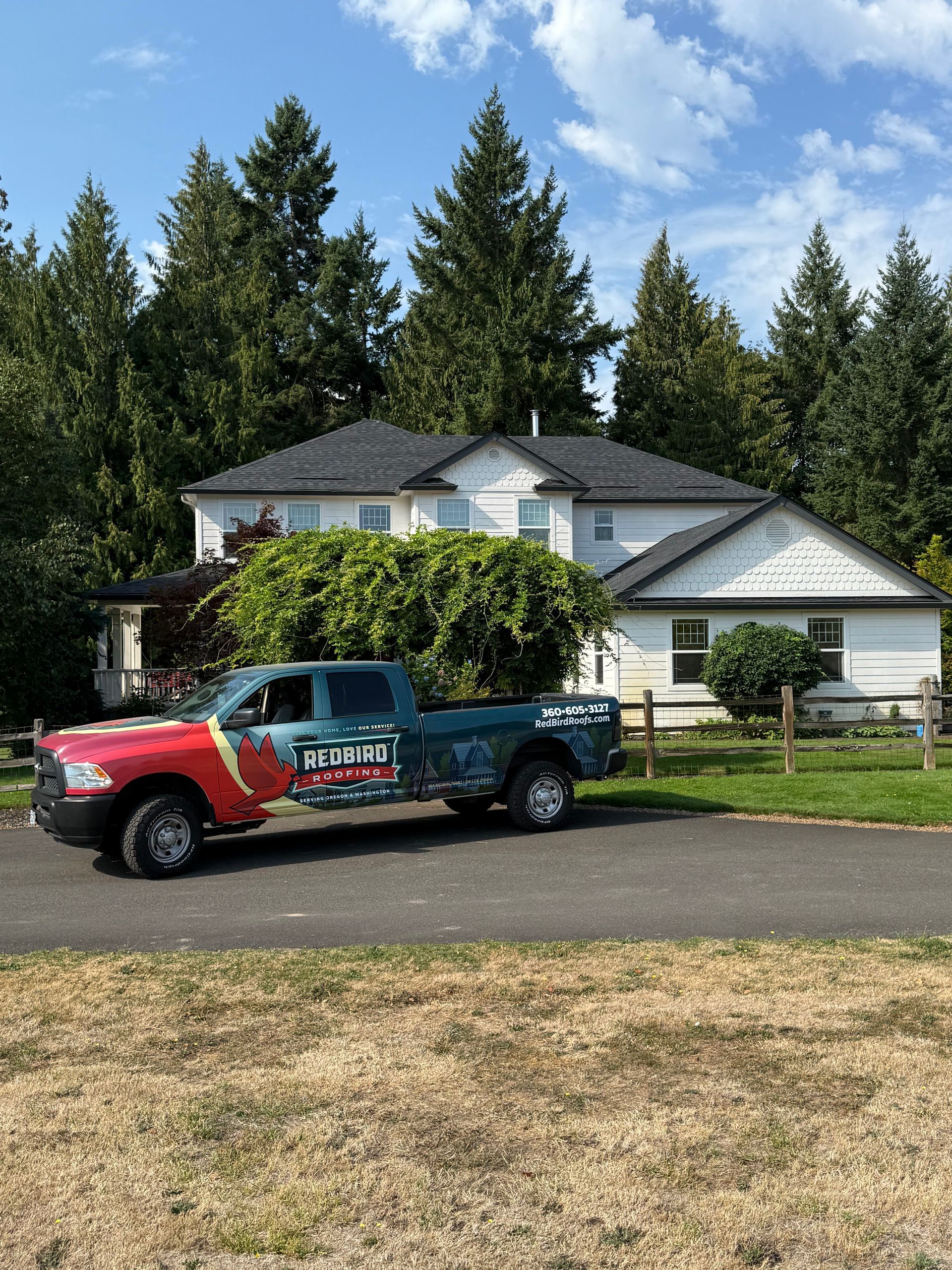 A work truck with a red front fender parked on a gravel driveway in front of a white two-story house surrounded by trees.