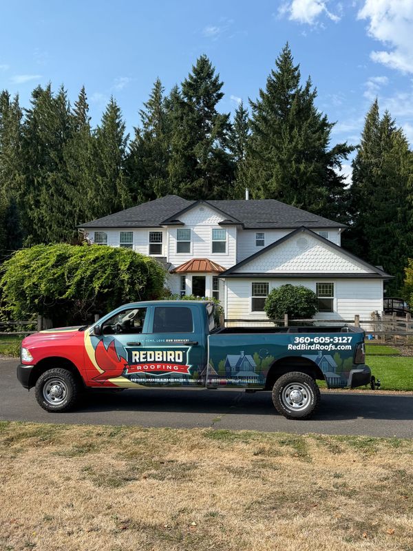 A teal and red work truck parked in front of a white two-story house with evergreen trees in the background.