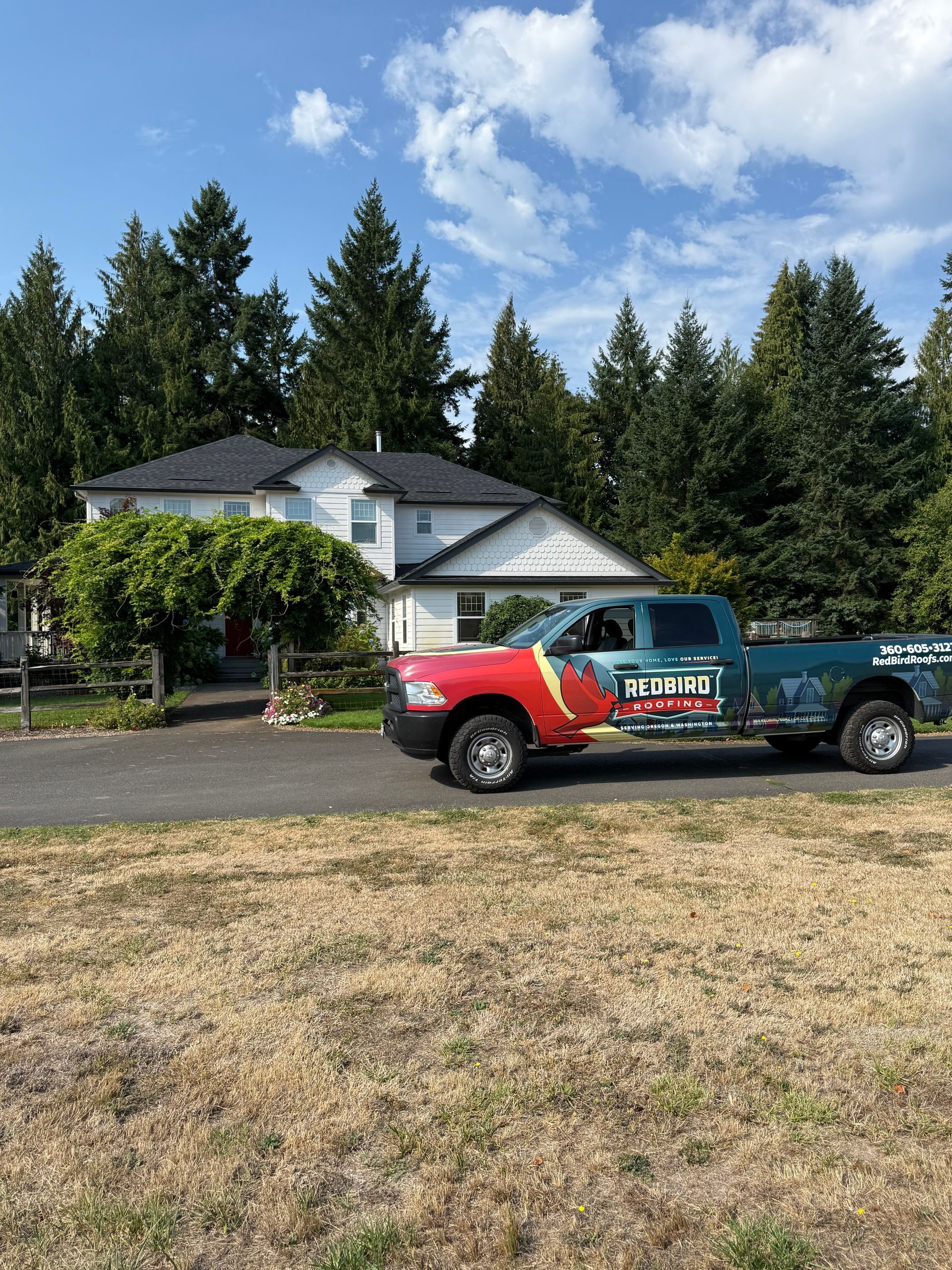 A multi-colored work truck is parked in front of a two-story house surrounded by evergreen trees on a sunny day.