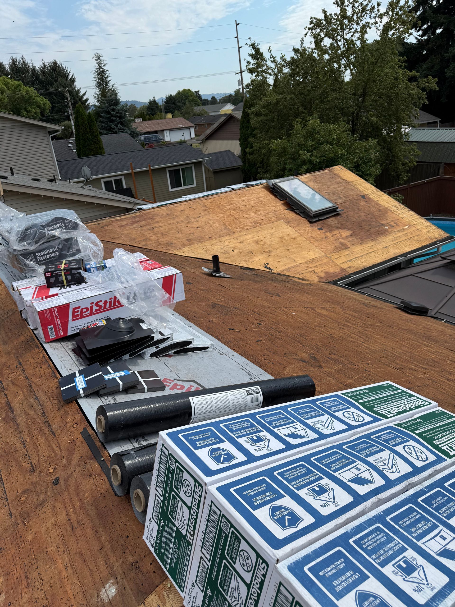View from a roof showing construction supplies, stacks of shingles, and a skylight on an adjacent bare roof section.