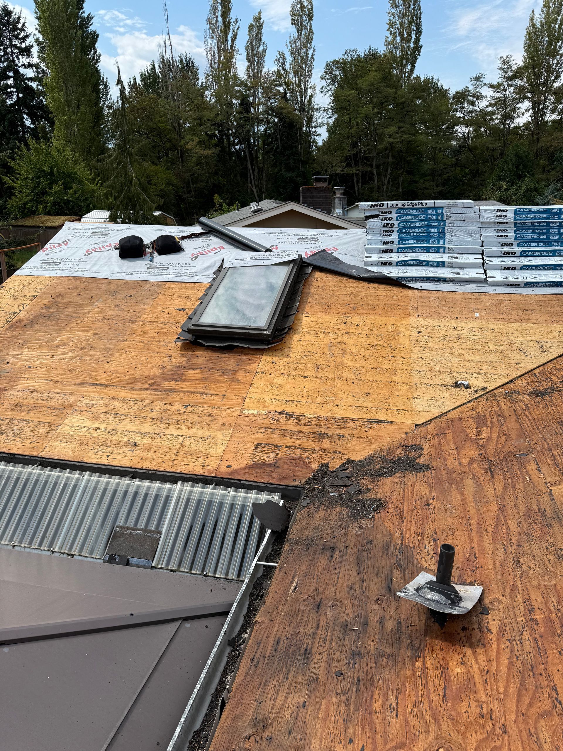 A roof under construction featuring exposed wooden sheathing, a skylight, a plumbing vent, and sections of underlayment.