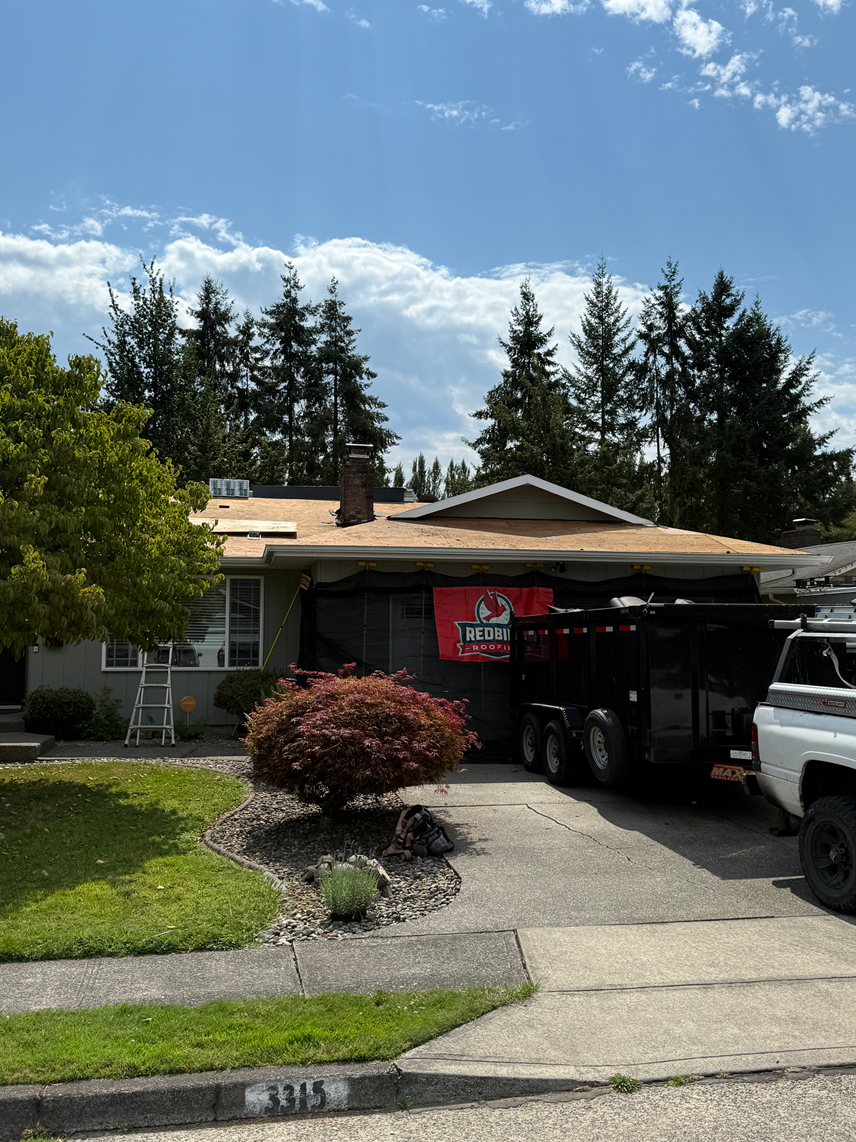 A one-story suburban house with a brown roof under repair, a large black dump trailer in the driveway, and tall pines.