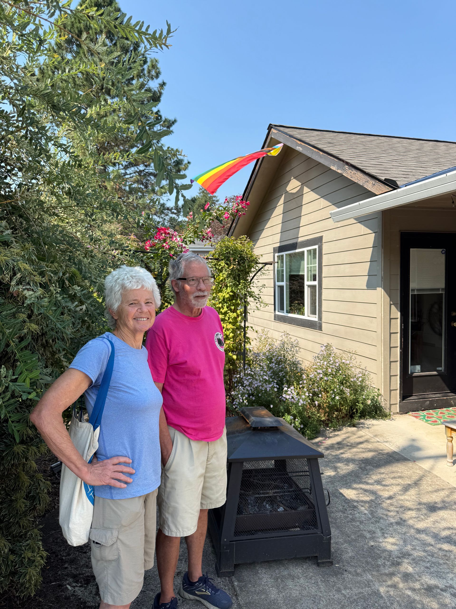 Two people stand together smiling outside a light-colored house with a rainbow-colored streamer attached to the roof.