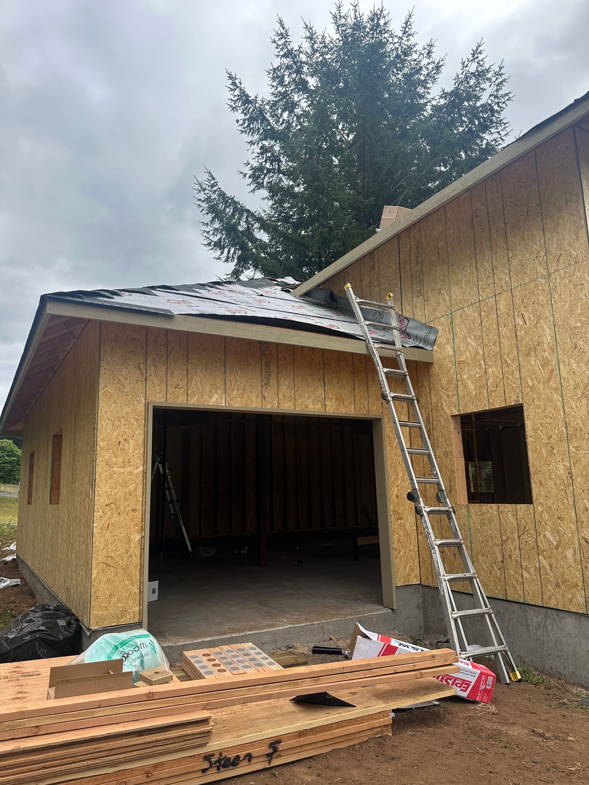 A garage under construction with plywood walls, a partially shingled roof, and a ladder leaning against the side.