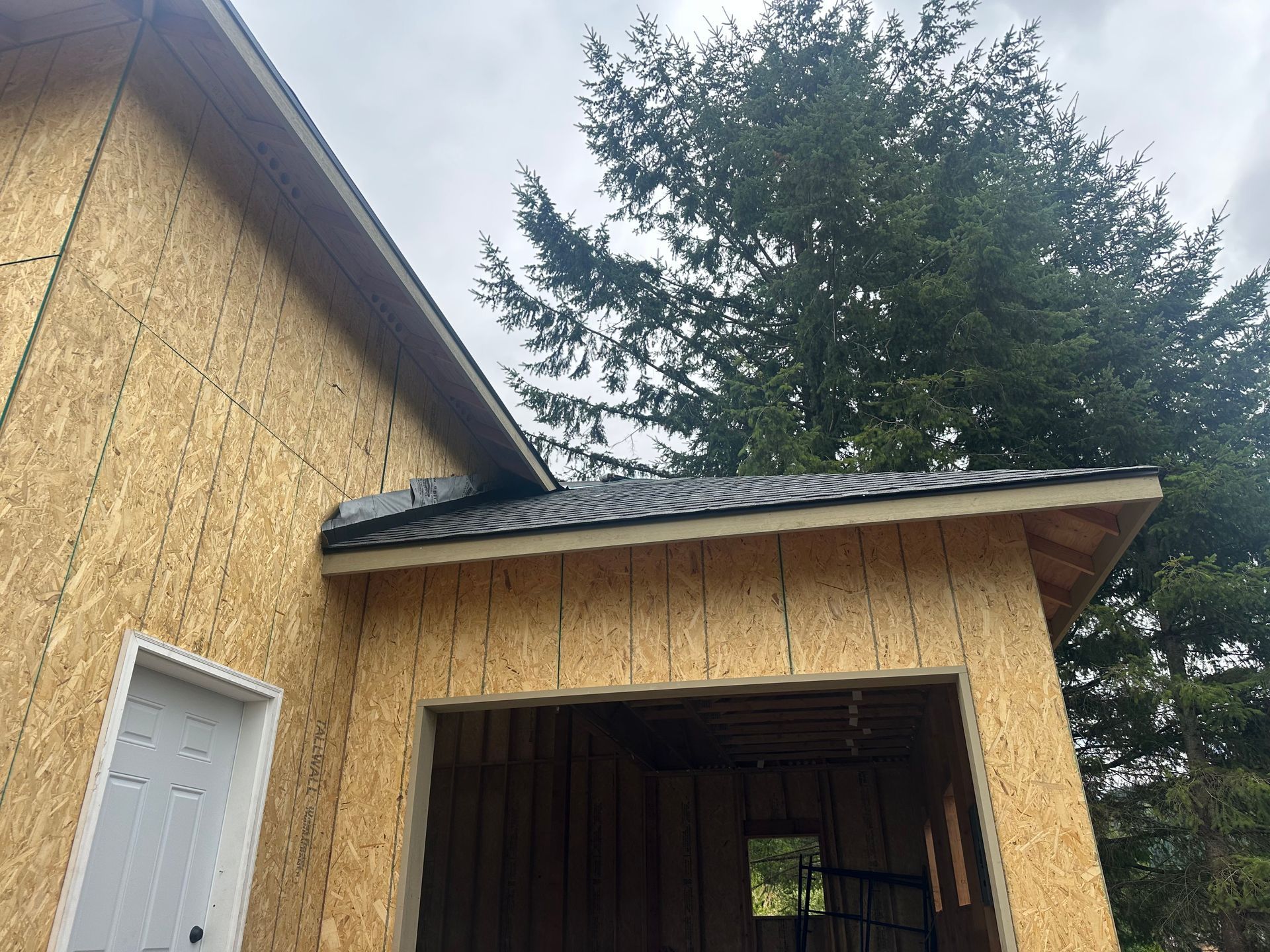 A newly framed garage exterior under construction, featuring oriented strand board walls and a partially shingled roof.