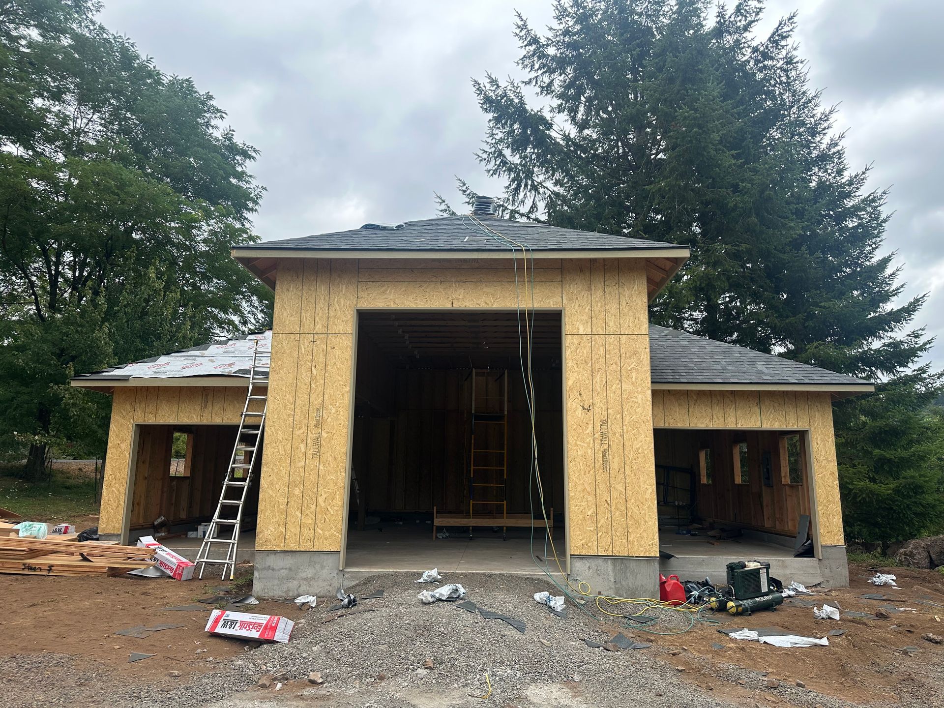 A new garage structure under construction with wood-sheathed walls, a shingled roof, and three open bays in a wooded lot.