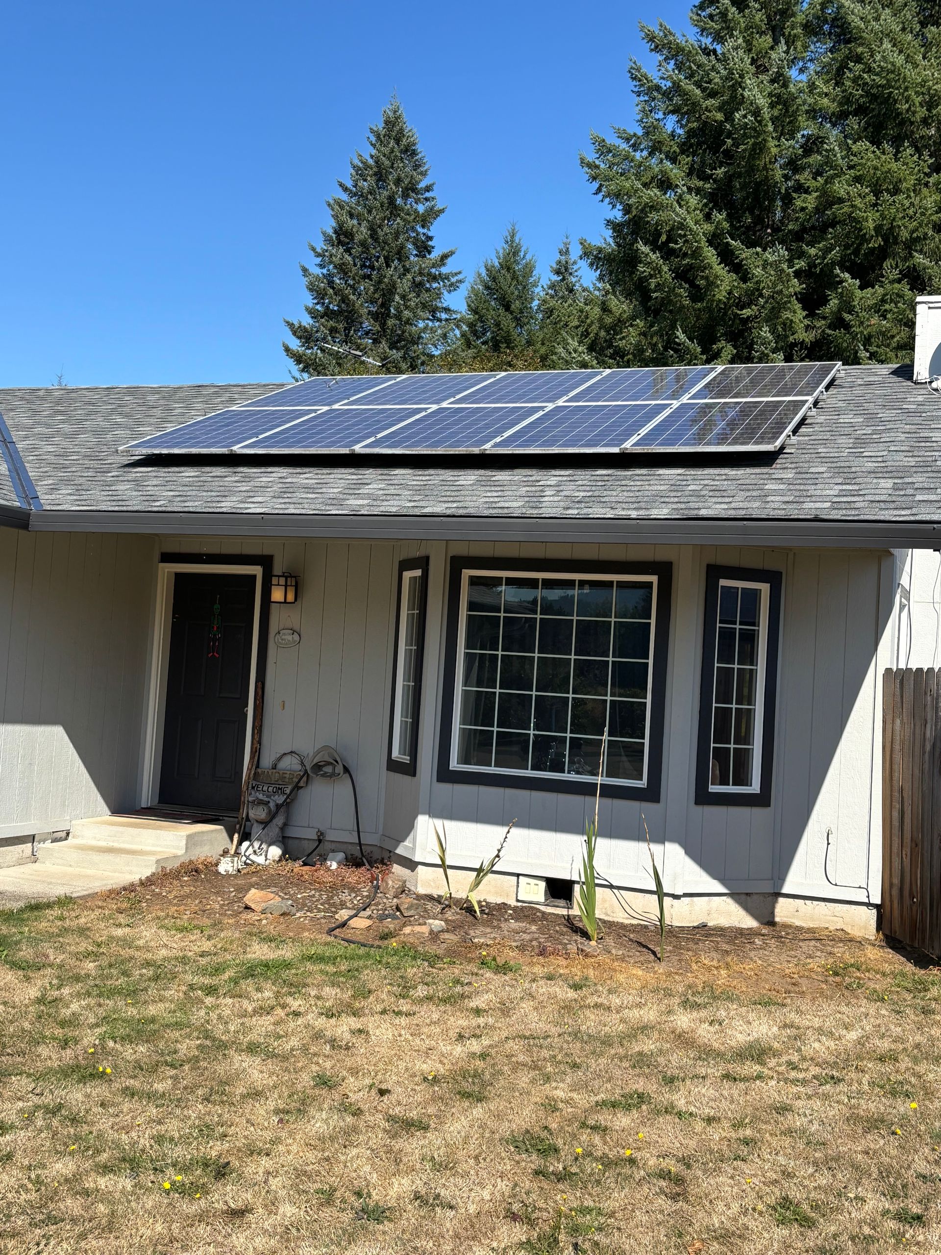 A single-story house with a grey roof featuring solar panels, a dark front door, and large windows, set against blue sky.