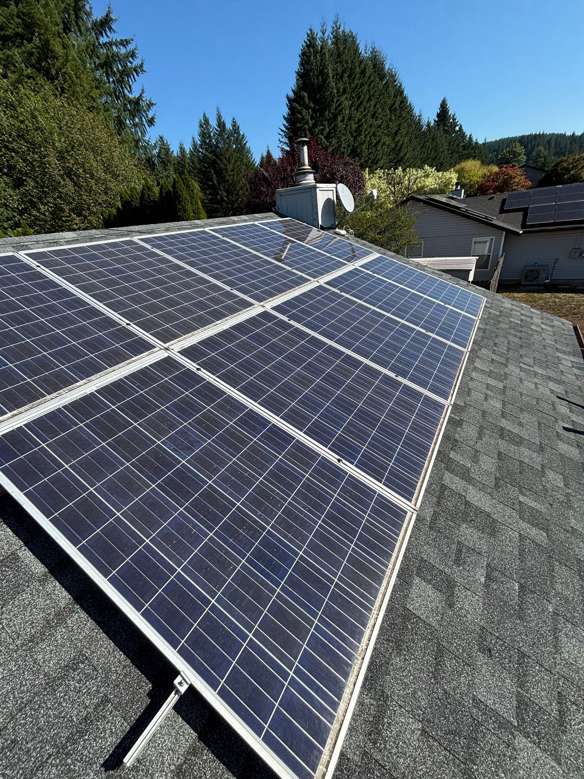 A solar panel array mounted on a gray shingled roof under a clear blue sky, surrounded by trees.