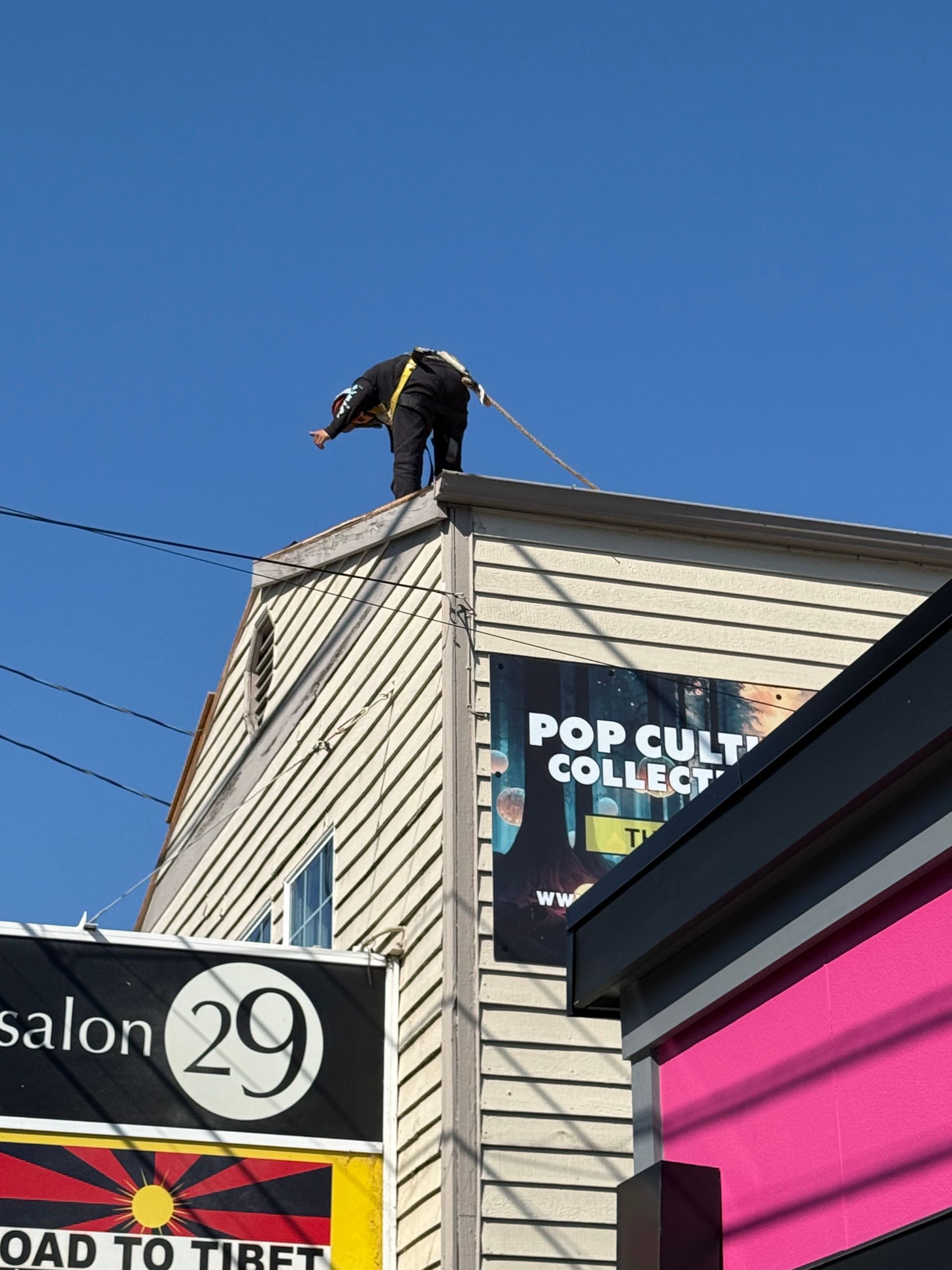 A person wearing safety gear stands on a rooftop, holding a rope against a clear blue sky above urban storefronts.