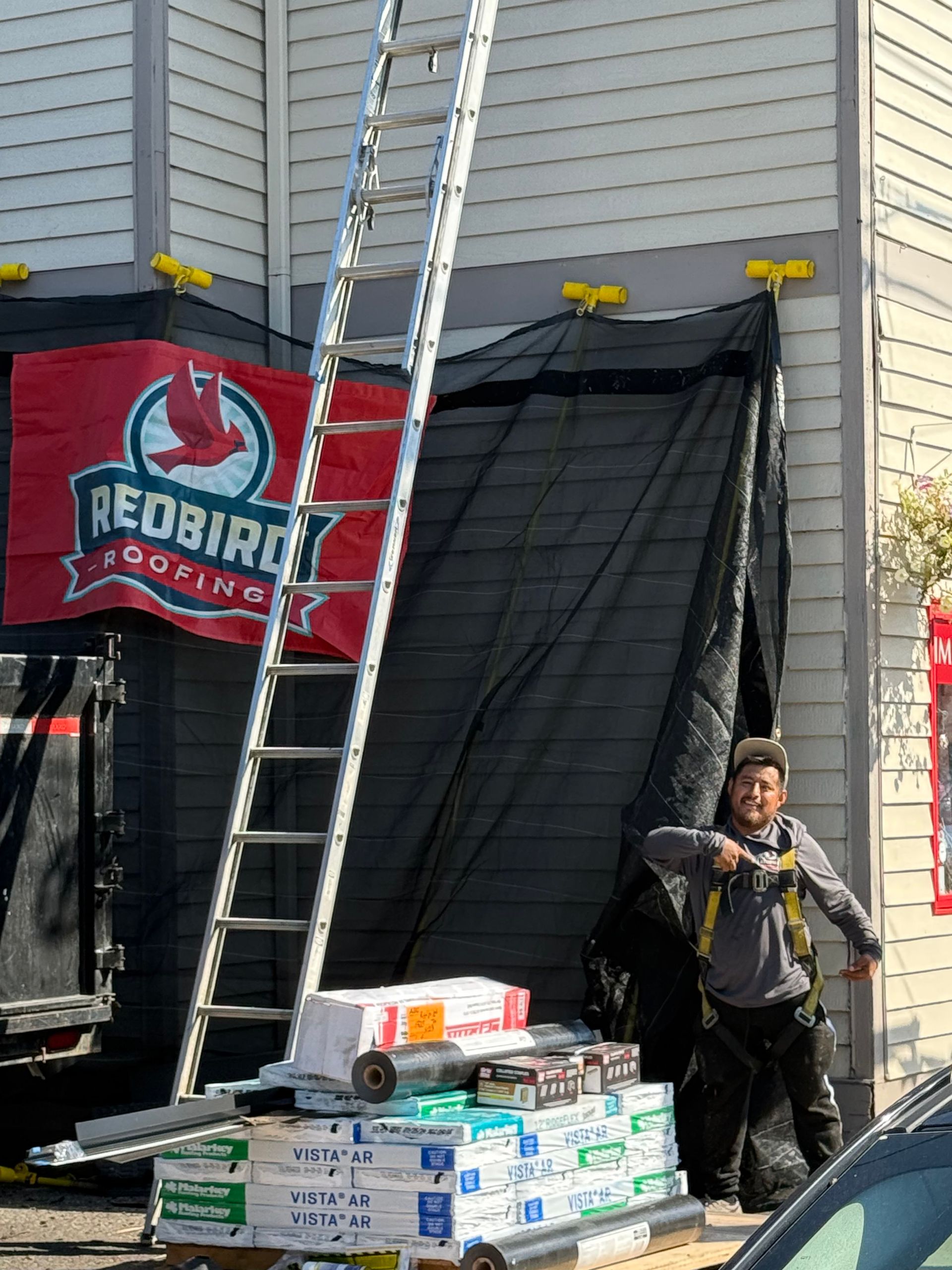 A worker in a safety harness stands near a ladder and stacked roofing supplies in front of a Redbird Roofing banner.