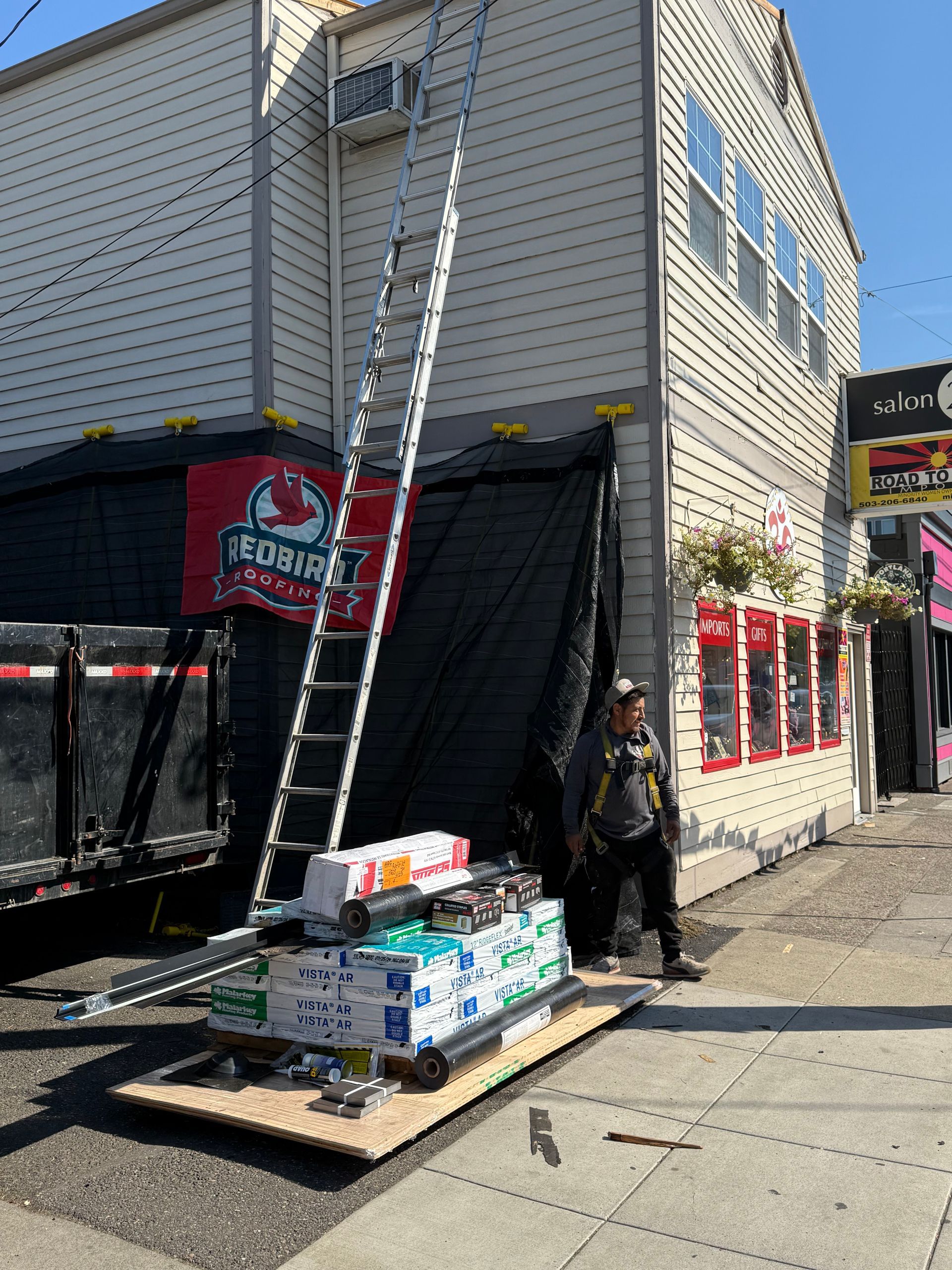 A construction worker stands beside a ladder leaning against a building, with a stack of supplies on a platform nearby.