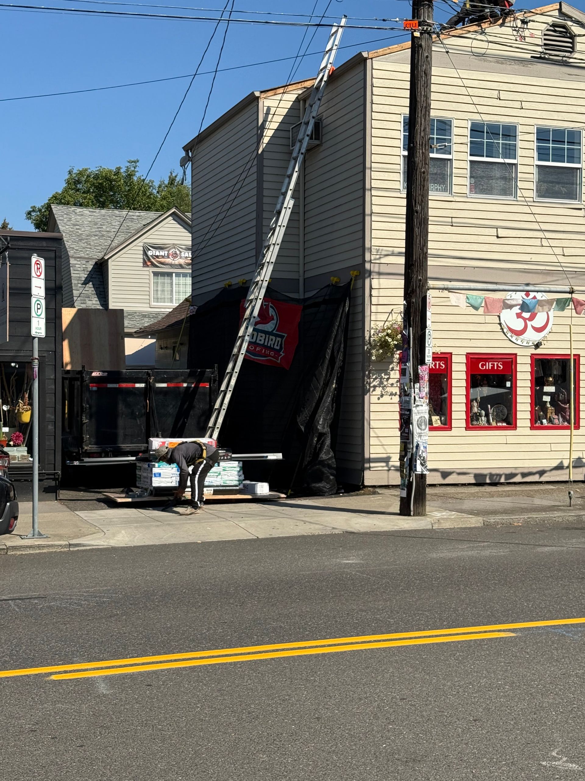 A long ladder is propped against the side of a tan two-story building near a utility pole and a work trailer.