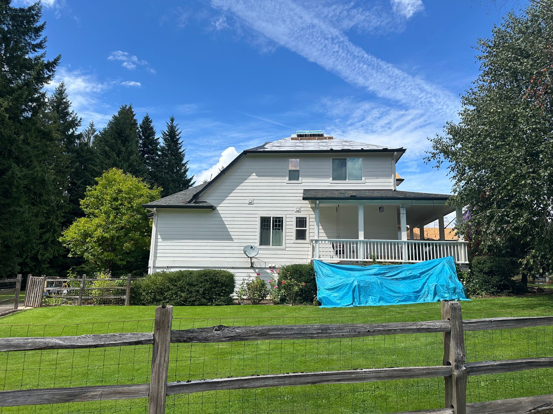 A white two-story house with a wooden porch and blue tarp stands in a green yard behind a rustic wooden fence.