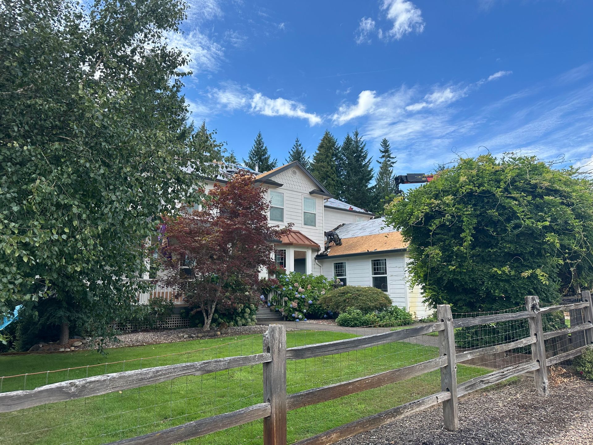 A two-story white house with a red-leaved tree in the front yard, seen behind a split-rail wooden fence on a sunny day.