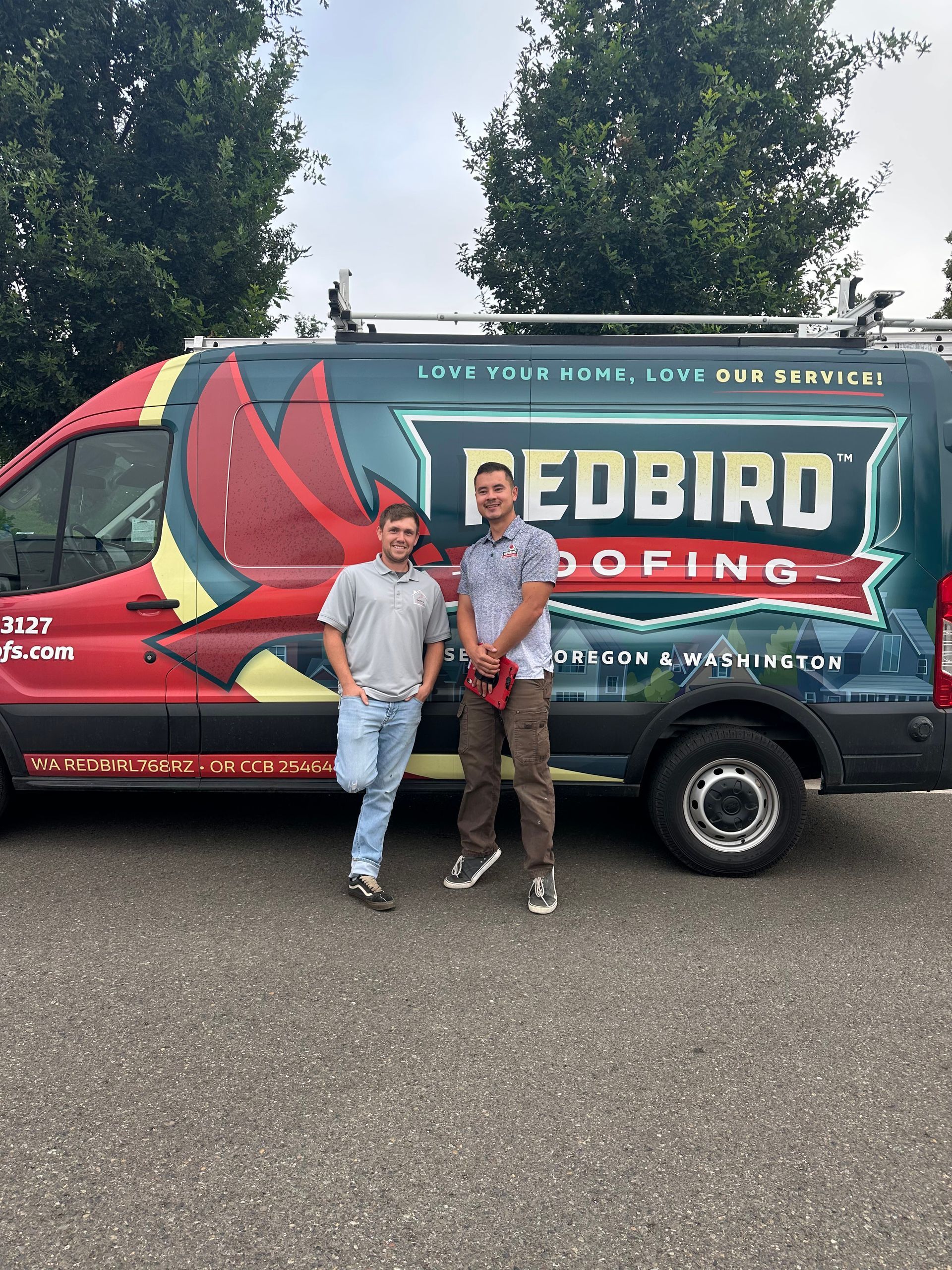 Two people stand in front of a Redbird Roofing van parked on asphalt.